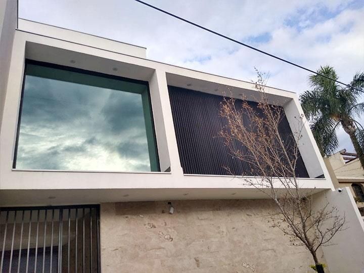 Edificio moderno de color blanco con un gran ventanal que refleja un cielo nublado y un panel de celosía oscura.