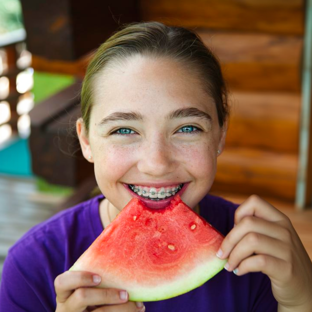 Smiling child holding a slice of watermelon outdoors, with braces visible and a wooden background