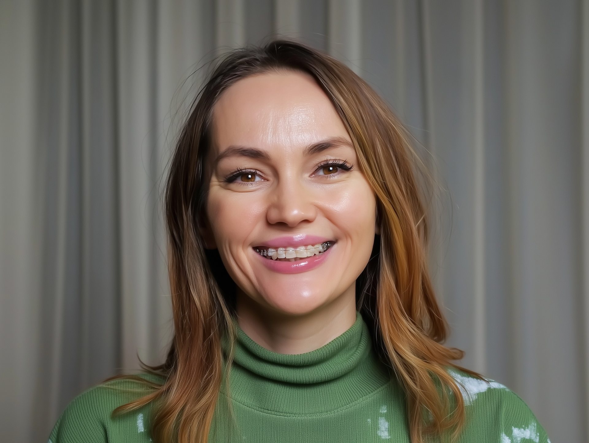 Woman with long brown hair smiles, wearing a green turtleneck sweater, against a beige curtain background.
