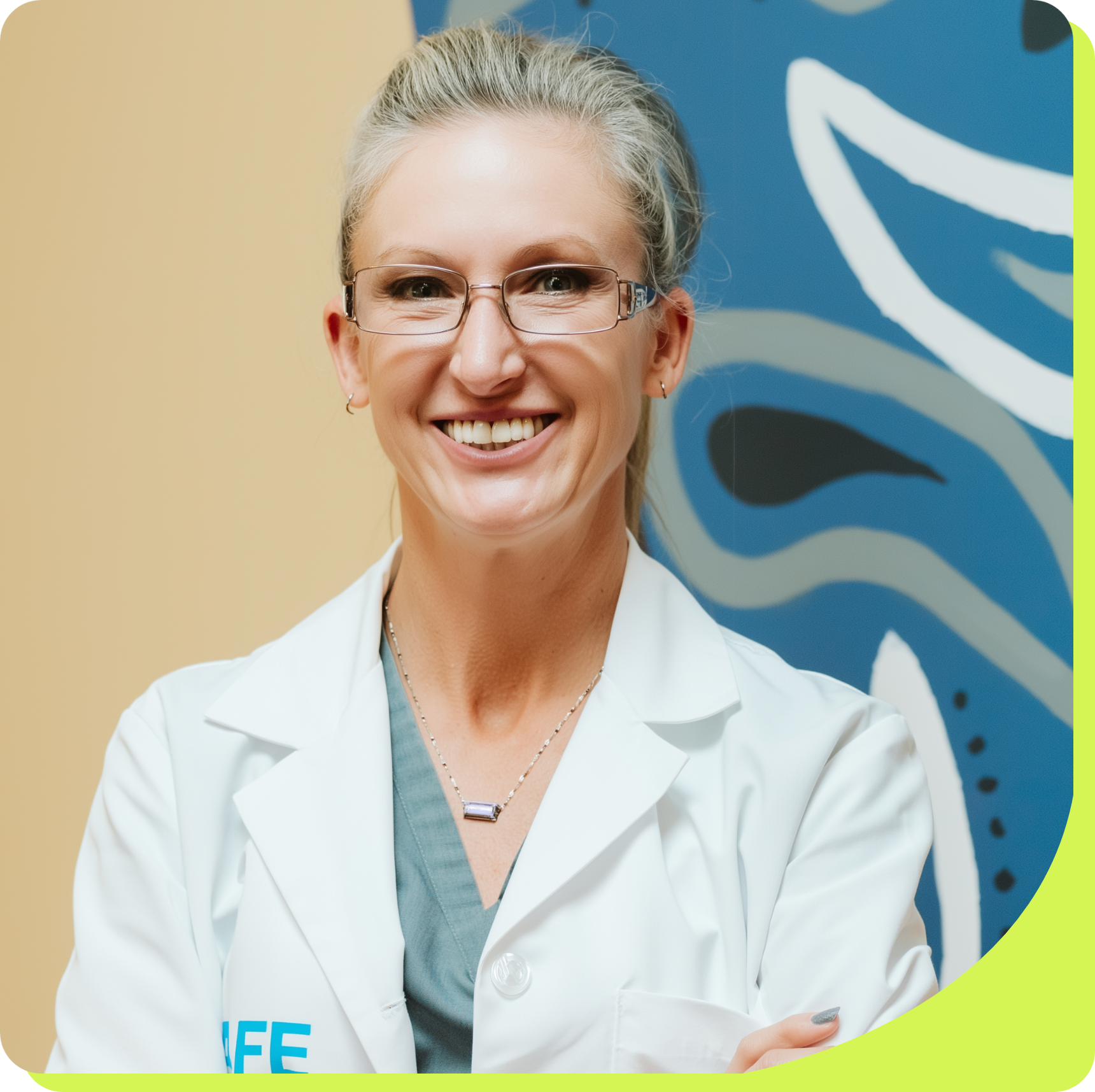 Woman in white coat, glasses, smiling, arms crossed, standing near a wall with blue and gray design.