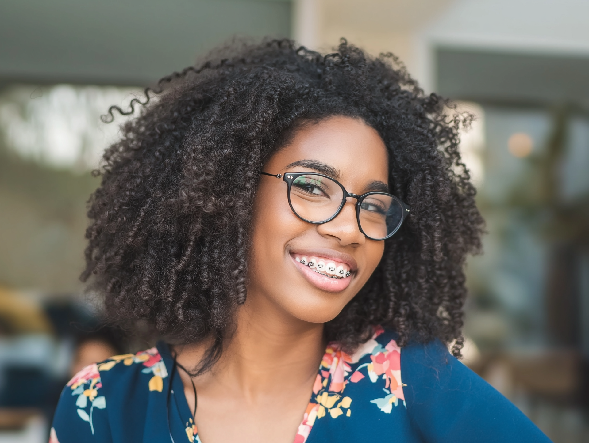 Person with curly hair, glasses, and braces smiles. Wearing a floral top. Outdoors.