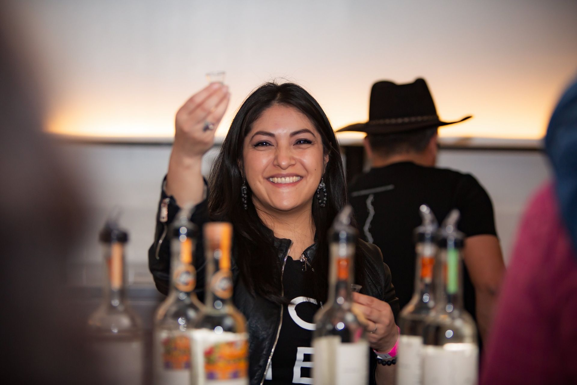 Mujer sonriente, sosteniendo un vaso de chupito, detrás de una barra con botellas de licor.