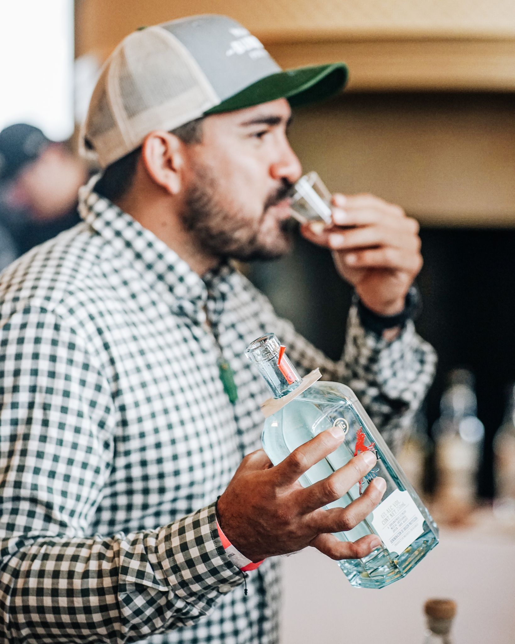 Hombre probando licor, sosteniendo una botella, vistiendo camisa a cuadros y gorra.