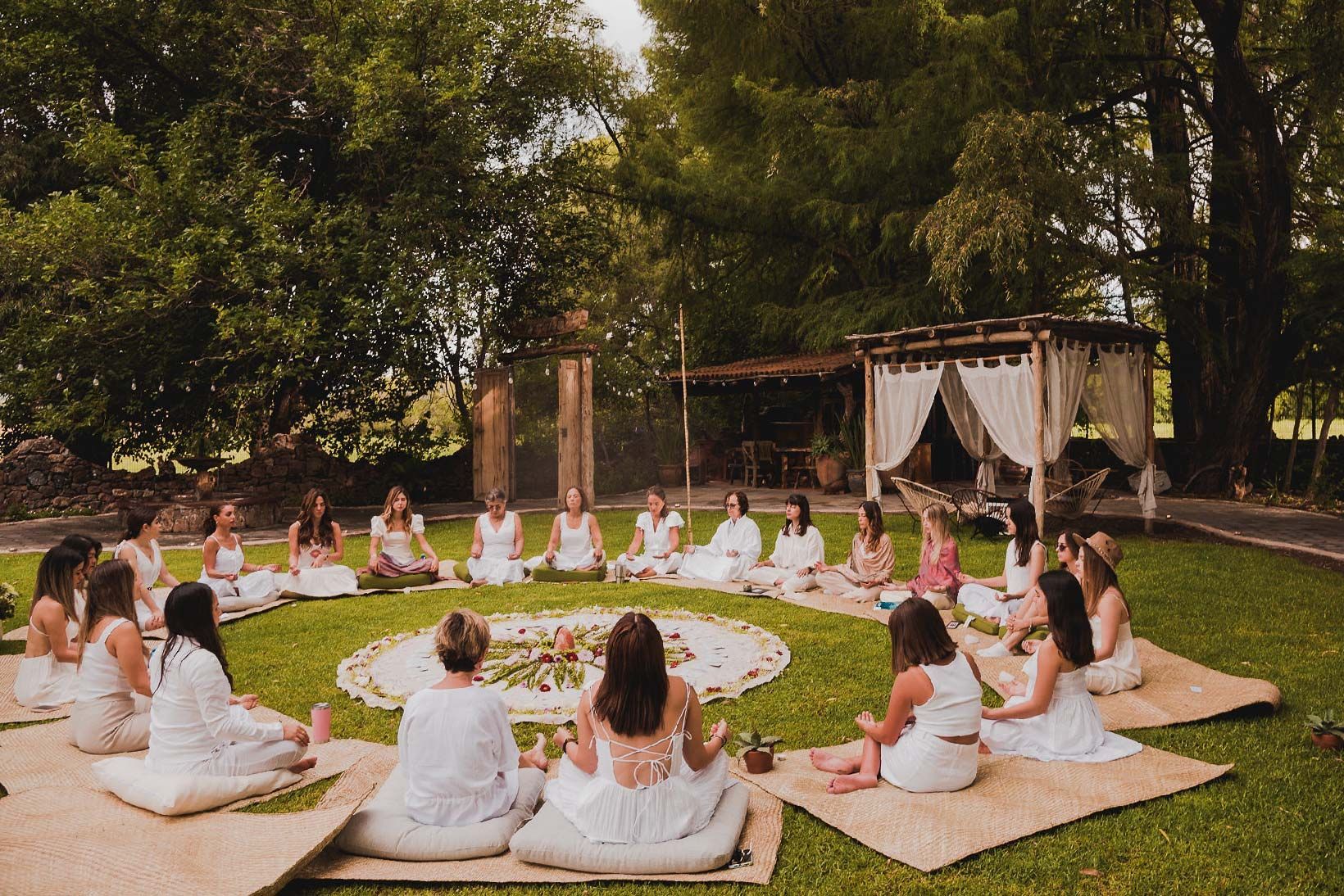Group of people in white clothing sitting in a circle on grass. Flowers and a draped structure are in the background.