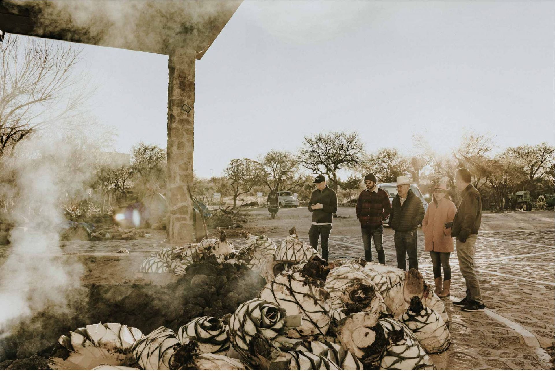 Personas observando el humo que se eleva de una pila de plantas de agave, al aire libre, con un edificio a la izquierda.