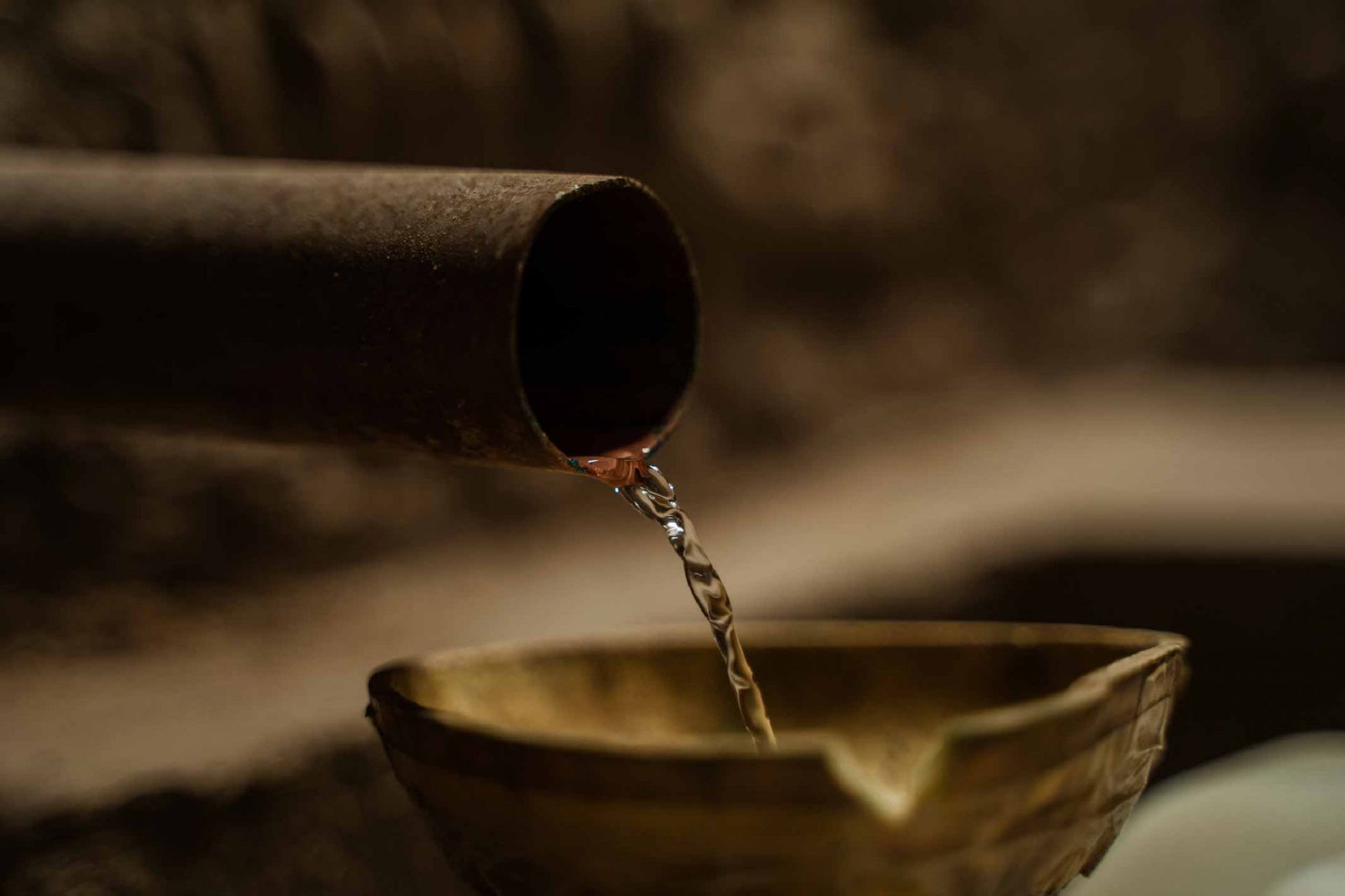 El agua fluye de una tubería oxidada a un cuenco de madera; ambiente interior.