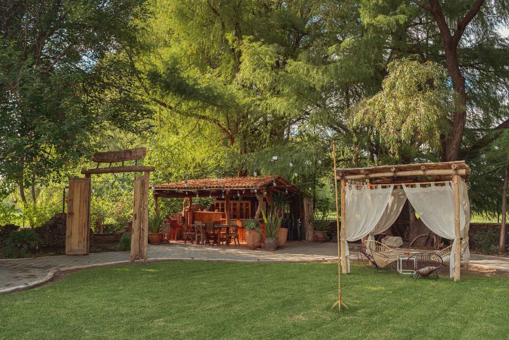 Lush green backyard with wooden structures: an open archway, a covered bar, and a draped bamboo canopy.