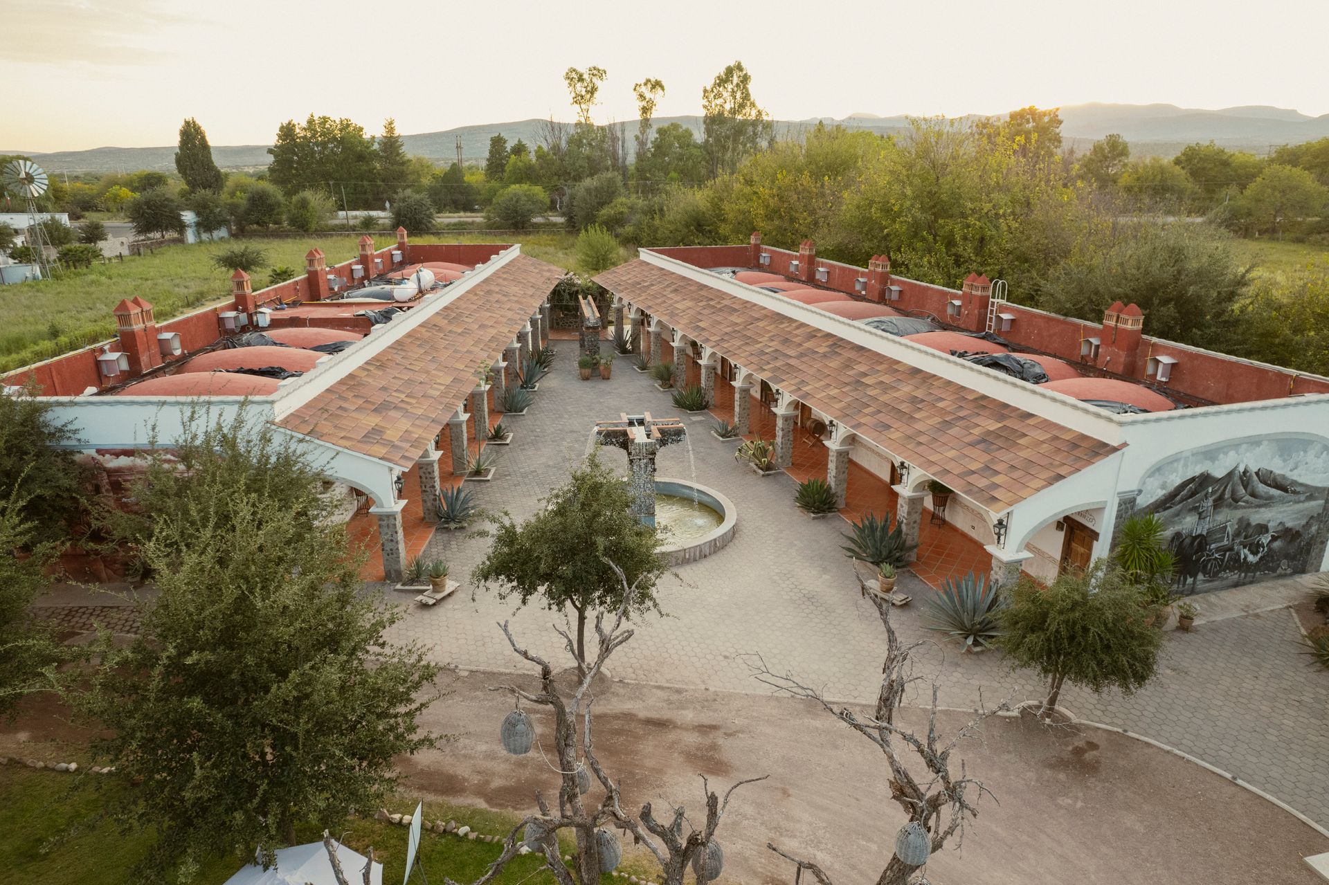 Dos edificios con tejados rojos se enfrentan entre sí, conectados por un patio con árboles, una fuente y un mural.