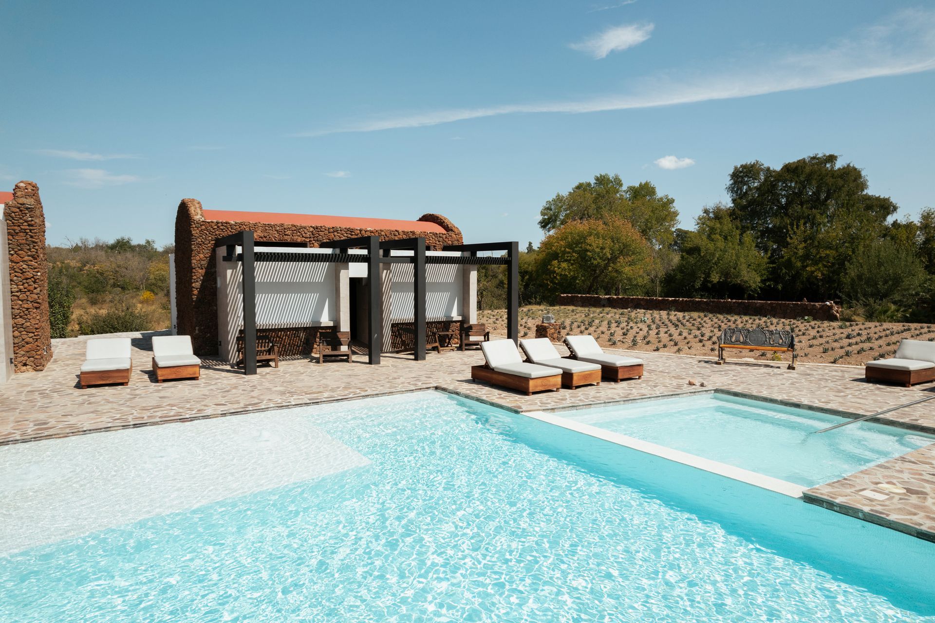 Zona de piscina con tumbonas, muros de piedra y una pequeña construcción con pérgola de madera bajo un cielo azul.