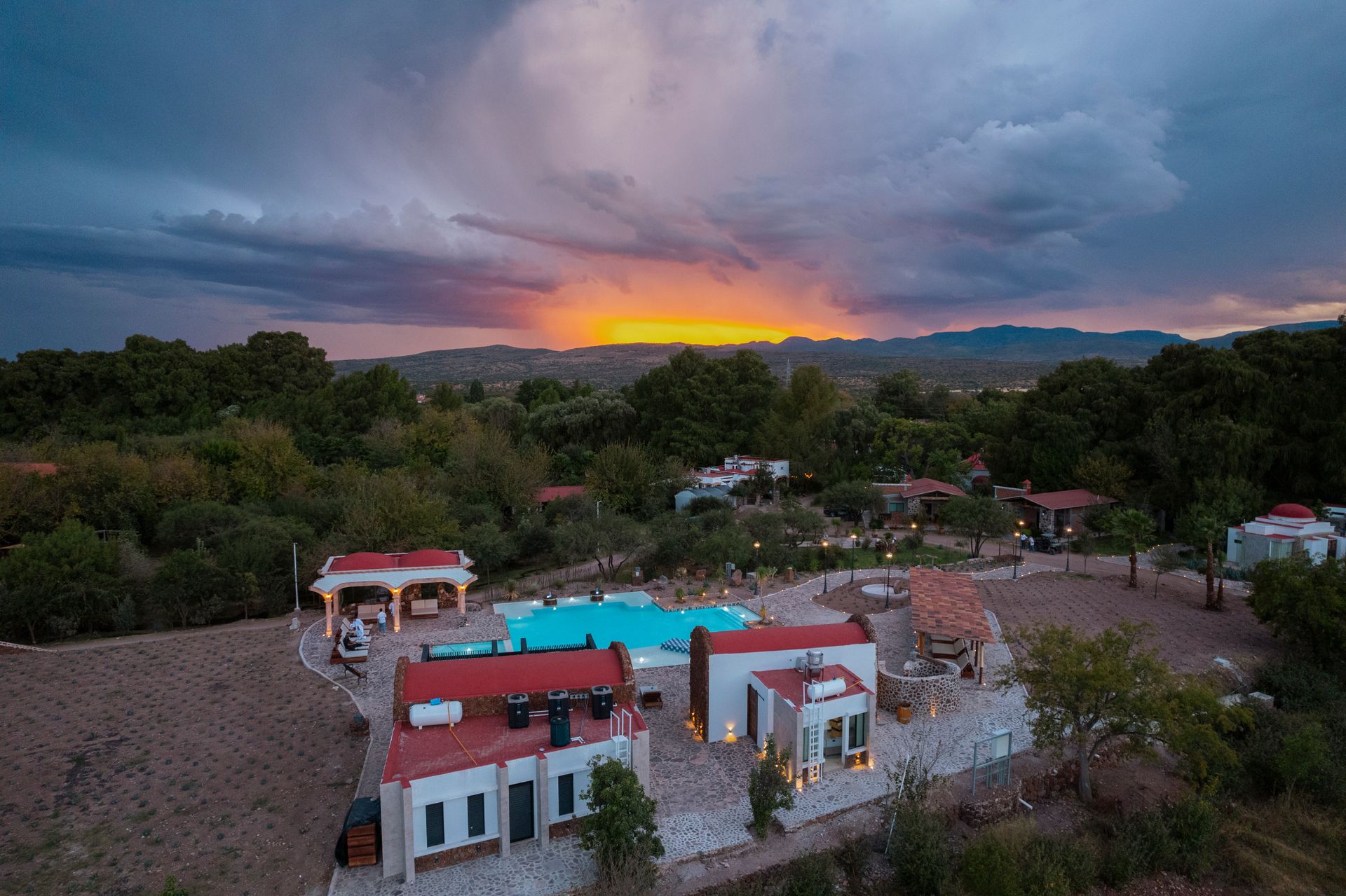 Vista del atardecer sobre un conjunto de edificios con piscina, rodeado de árboles.