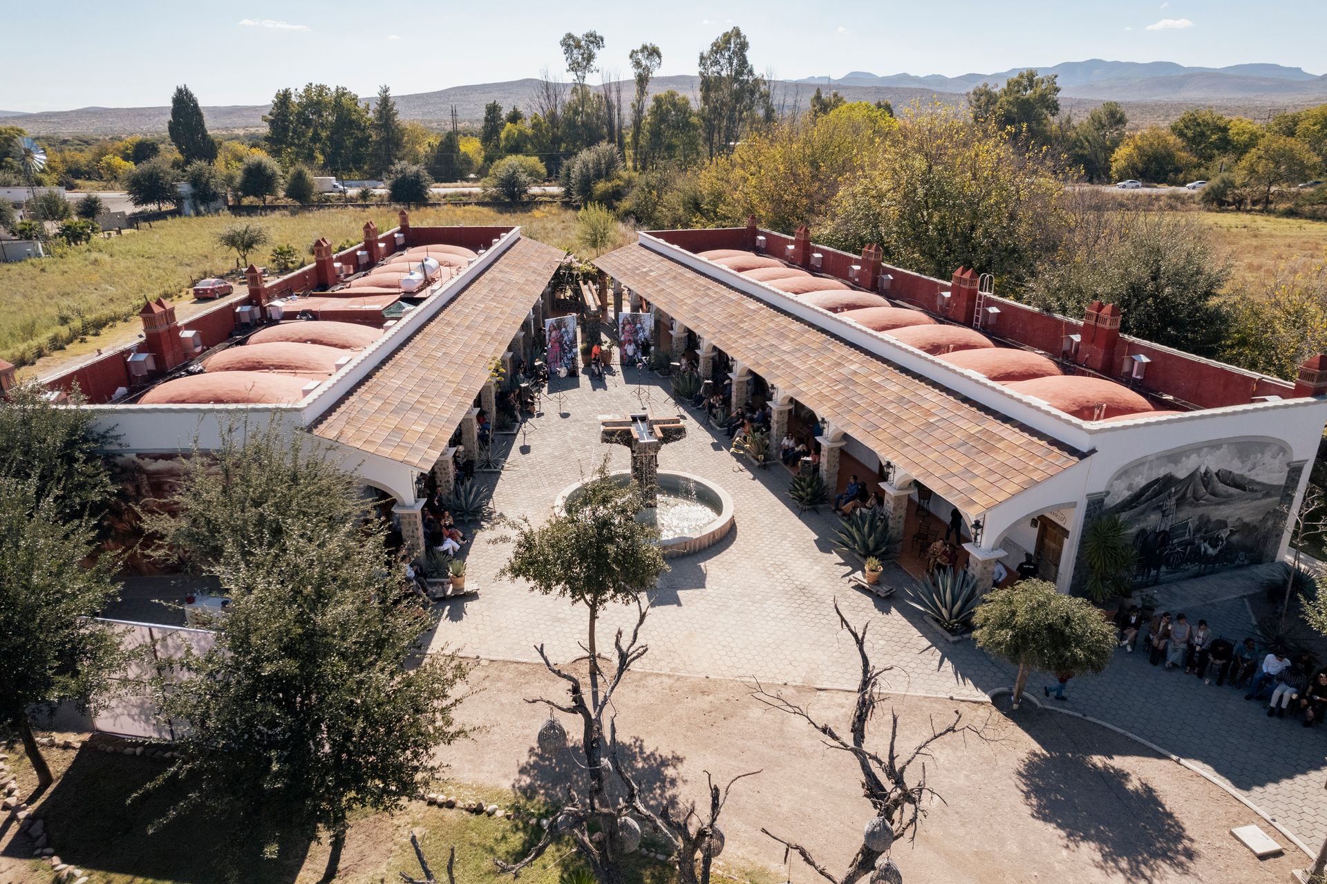 Courtyard with two long, red-roofed buildings. People gather in the center near a fountain. Trees surround the area.