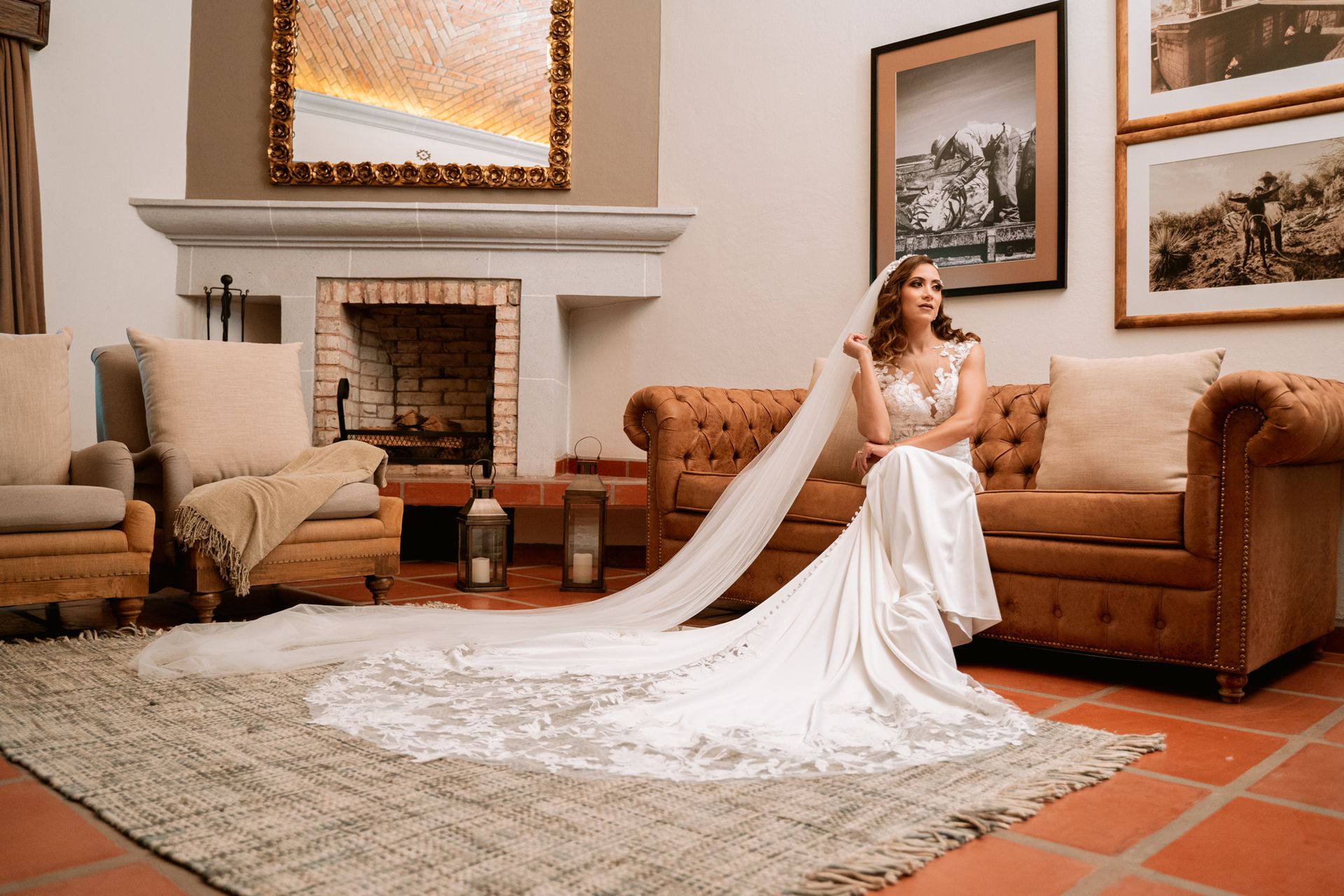 Bride in white gown with long train sits on a brown couch. She's in a room with a fireplace and artwork.
