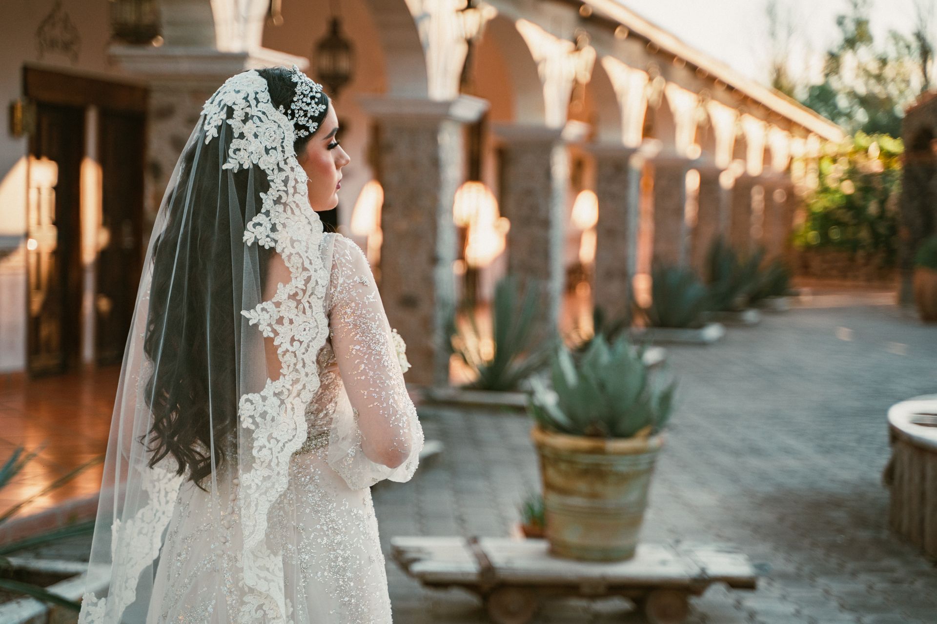 Bride in a sparkly gown and veil, stands outdoors, looking toward a stone colonnade with plants in terracotta pots.