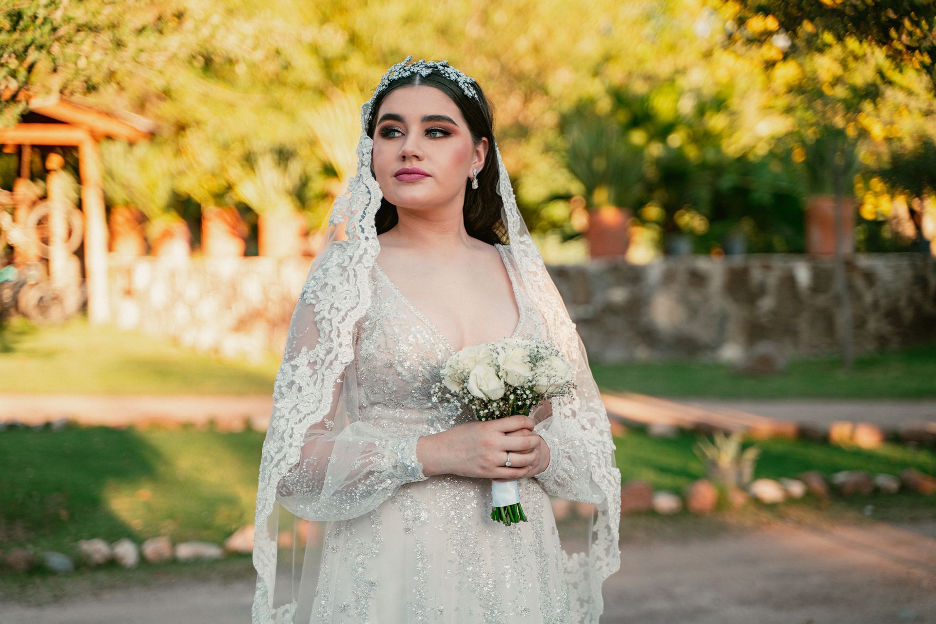 Bride in white gown, holding flowers, wearing veil and tiara, outdoors in soft sunlight.