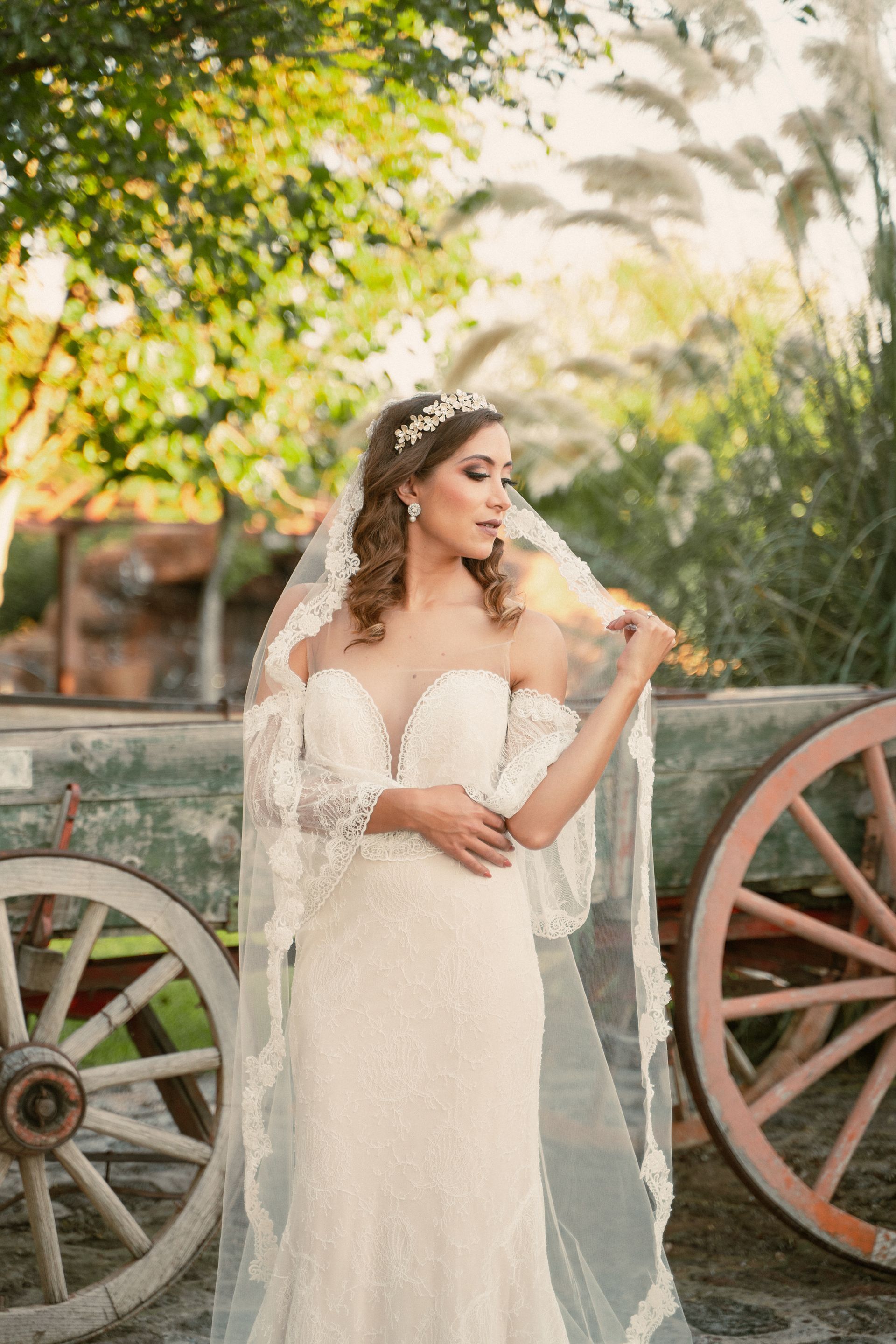 Bride in a white gown and veil poses outdoors near rustic wagon wheels and foliage.