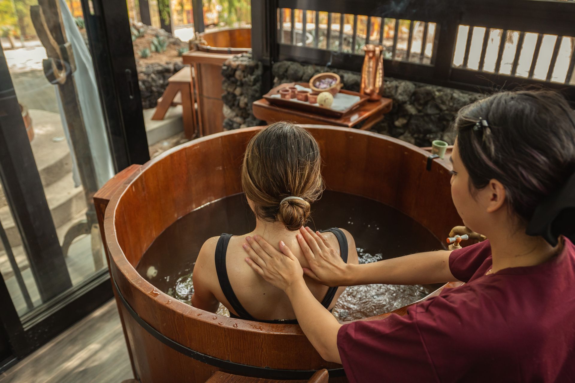 Mujer en una bañera de madera recibiendo un masaje de una persona con las manos en su espalda; ambiente de spa.