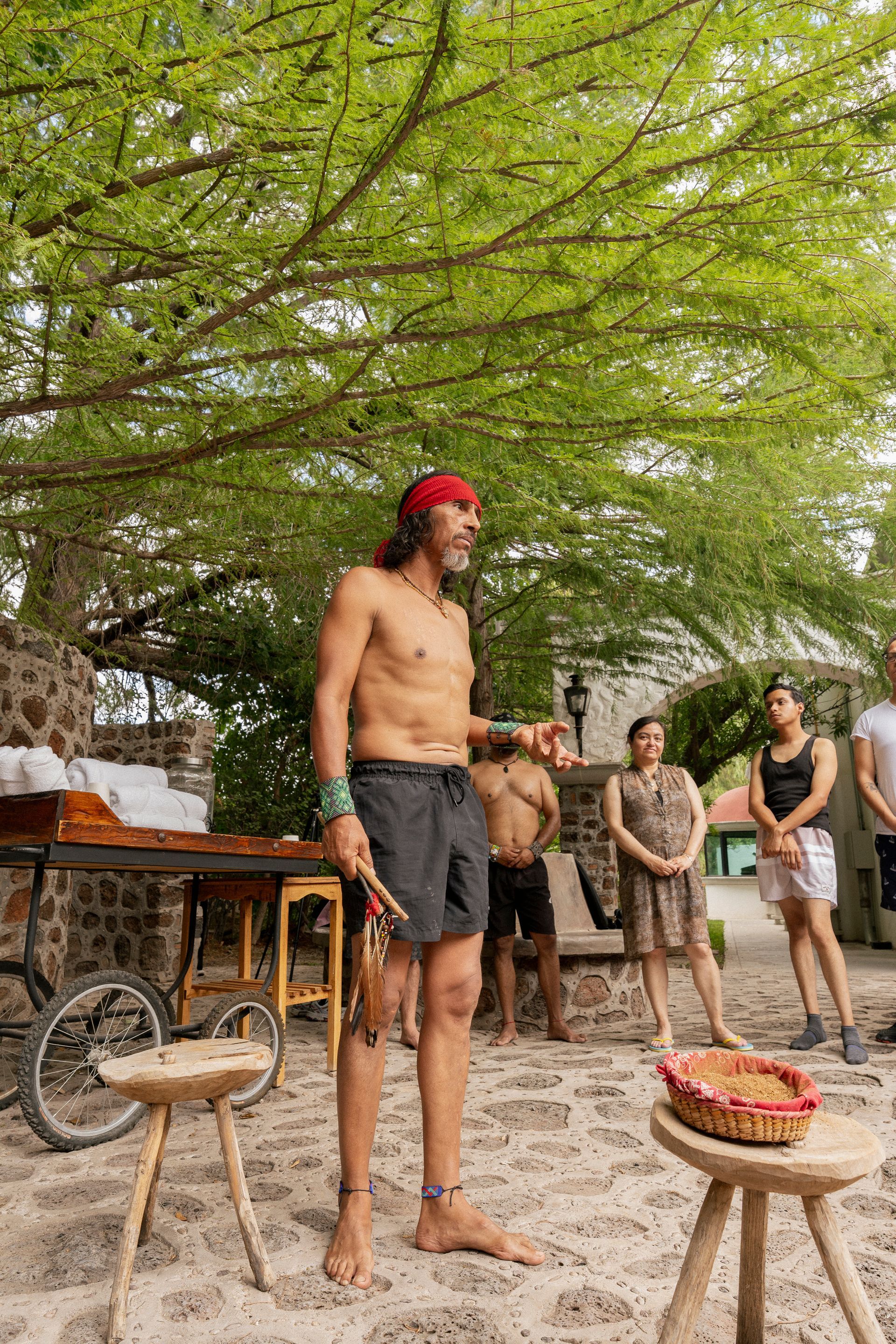 Un hombre con un pañuelo rojo habla, gesticulando con las manos hacia un grupo que se encuentra al aire libre.