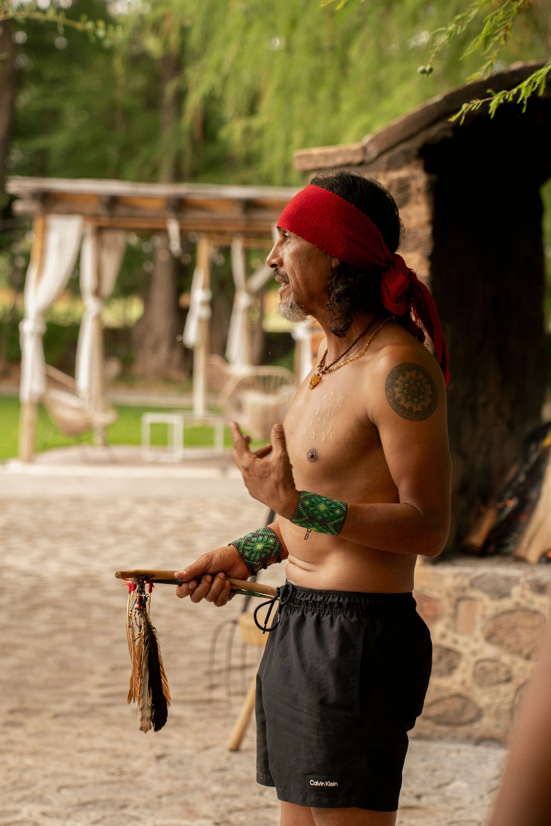 Hombre con cinta roja en la cabeza, sin camisa, gesticulando. Sostiene un objeto de madera con plumas. 
