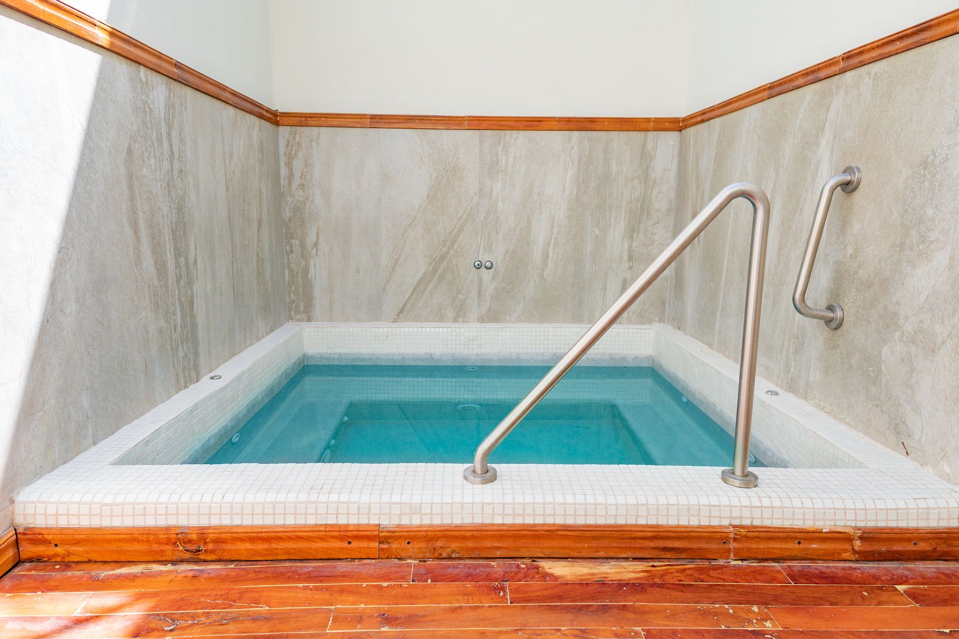 Un jacuzzi interior cuadrado con barandillas plateadas, rodeado de paredes de mármol, molduras de madera y suelo de madera.