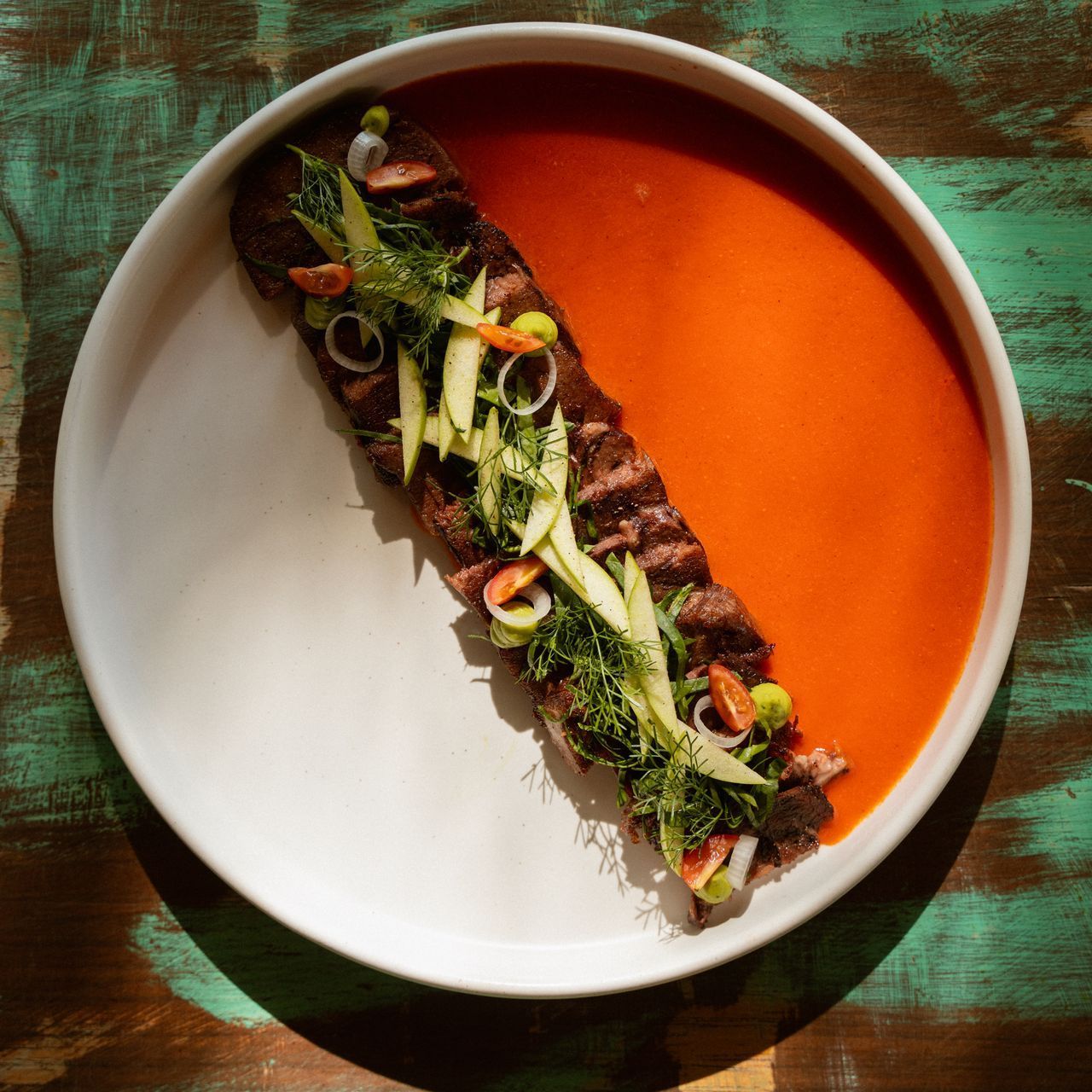 Plate with seared meat, herbs, and vegetables beside orange sauce.