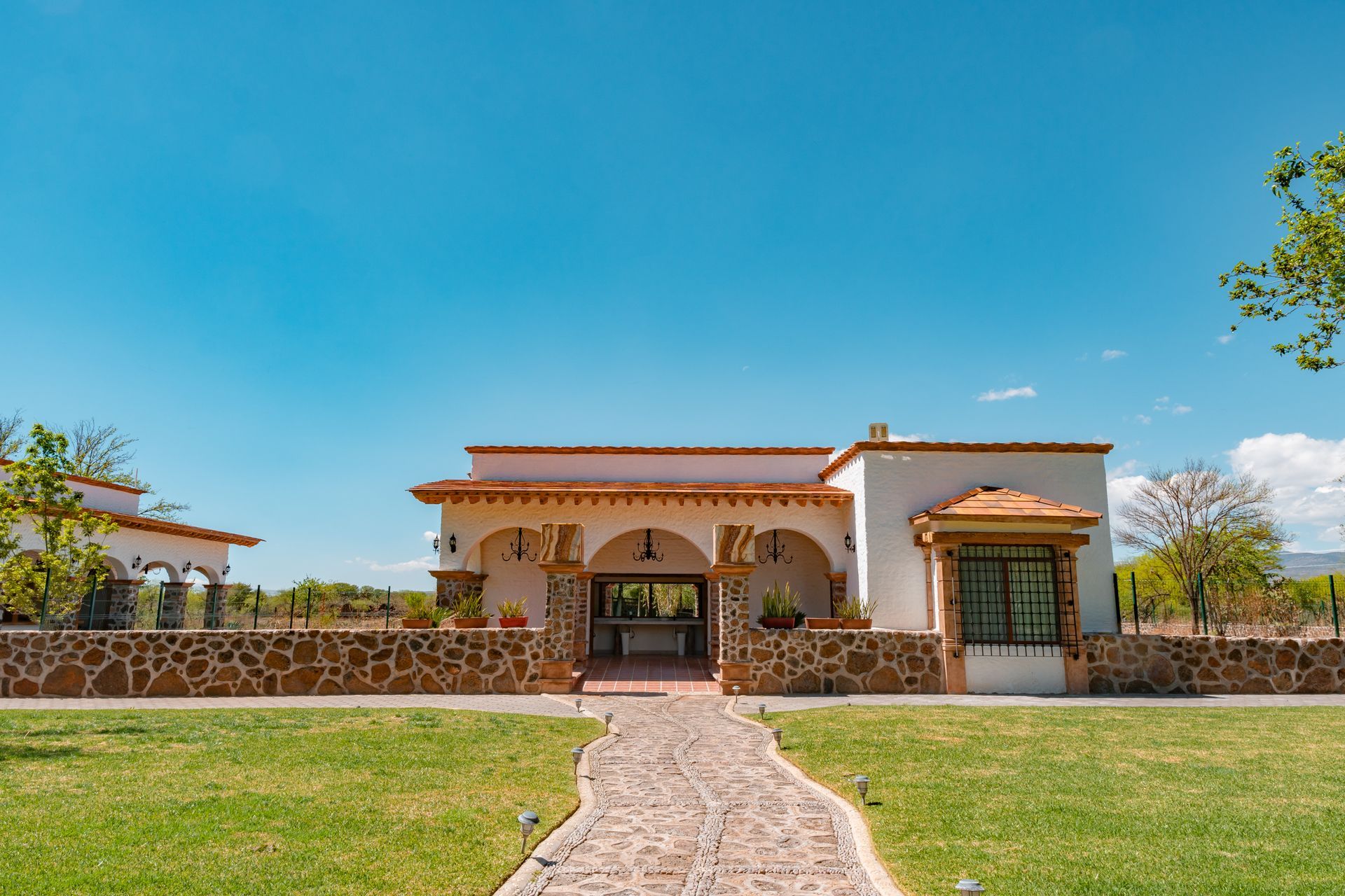 White stucco building with arched entryway, stone wall, and green lawn under a blue sky.