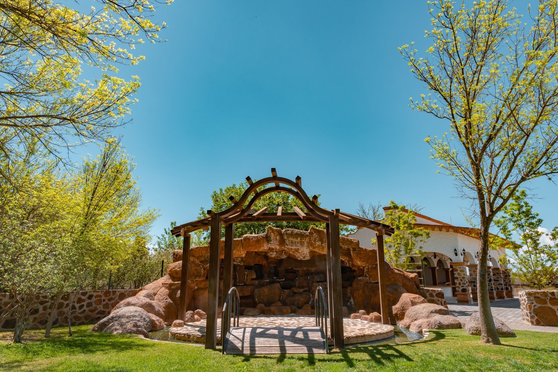 Wooden pergola over a rocky area with a building in the background, set under a blue sky and framed by trees.