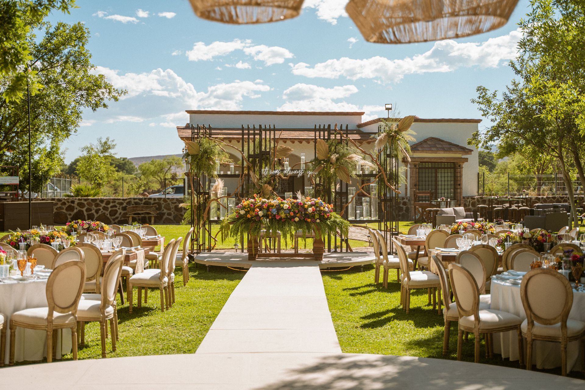 Wedding reception setup on a lawn; tables, chairs, floral decorations, and a decorated backdrop.