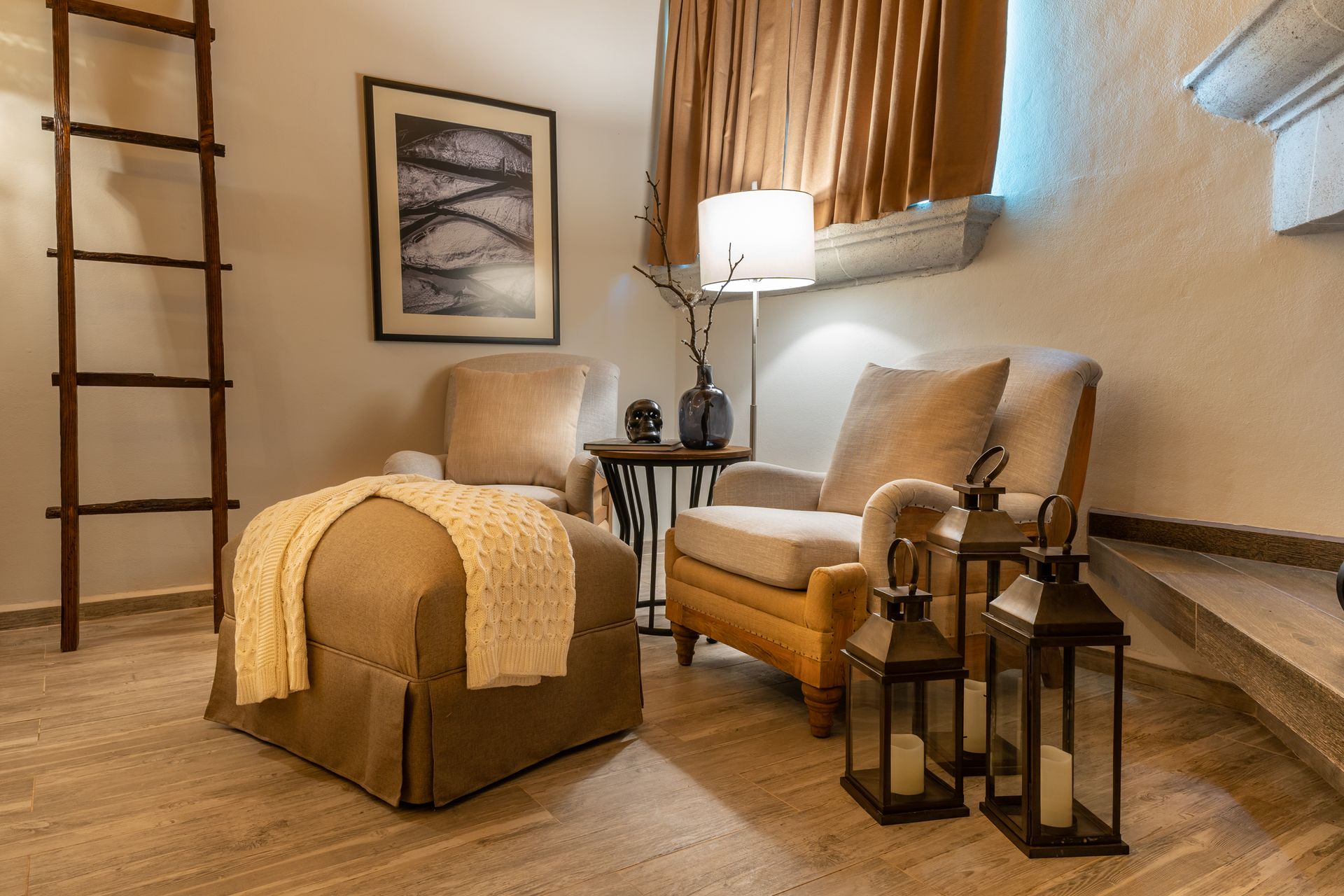 Cozy reading nook with two armchairs, ottoman, decorative lanterns, and ladder against a wall with a painting.
