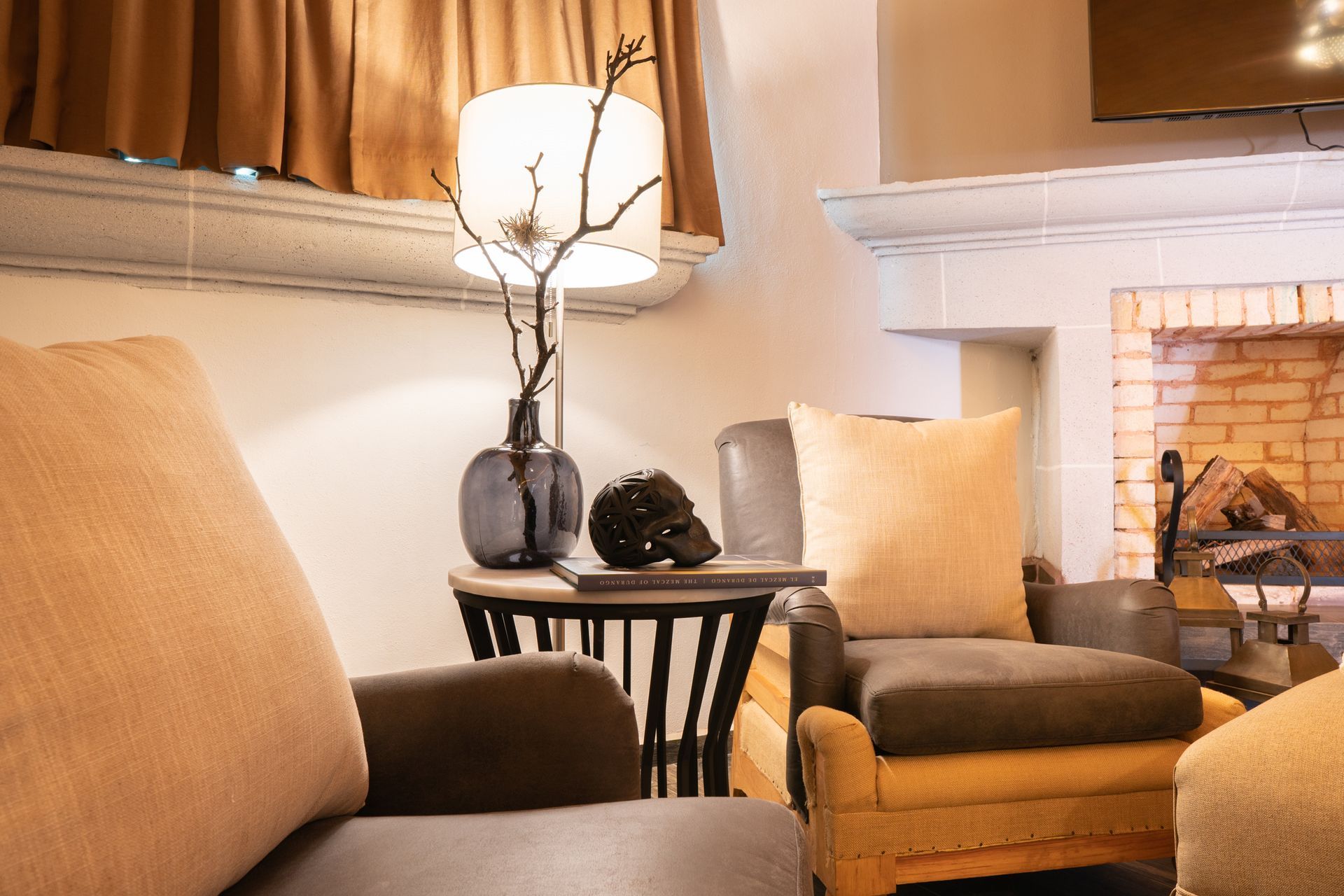 Cozy living room with armchairs, a side table with a skull, and a fireplace. Warm lighting and neutral colors.