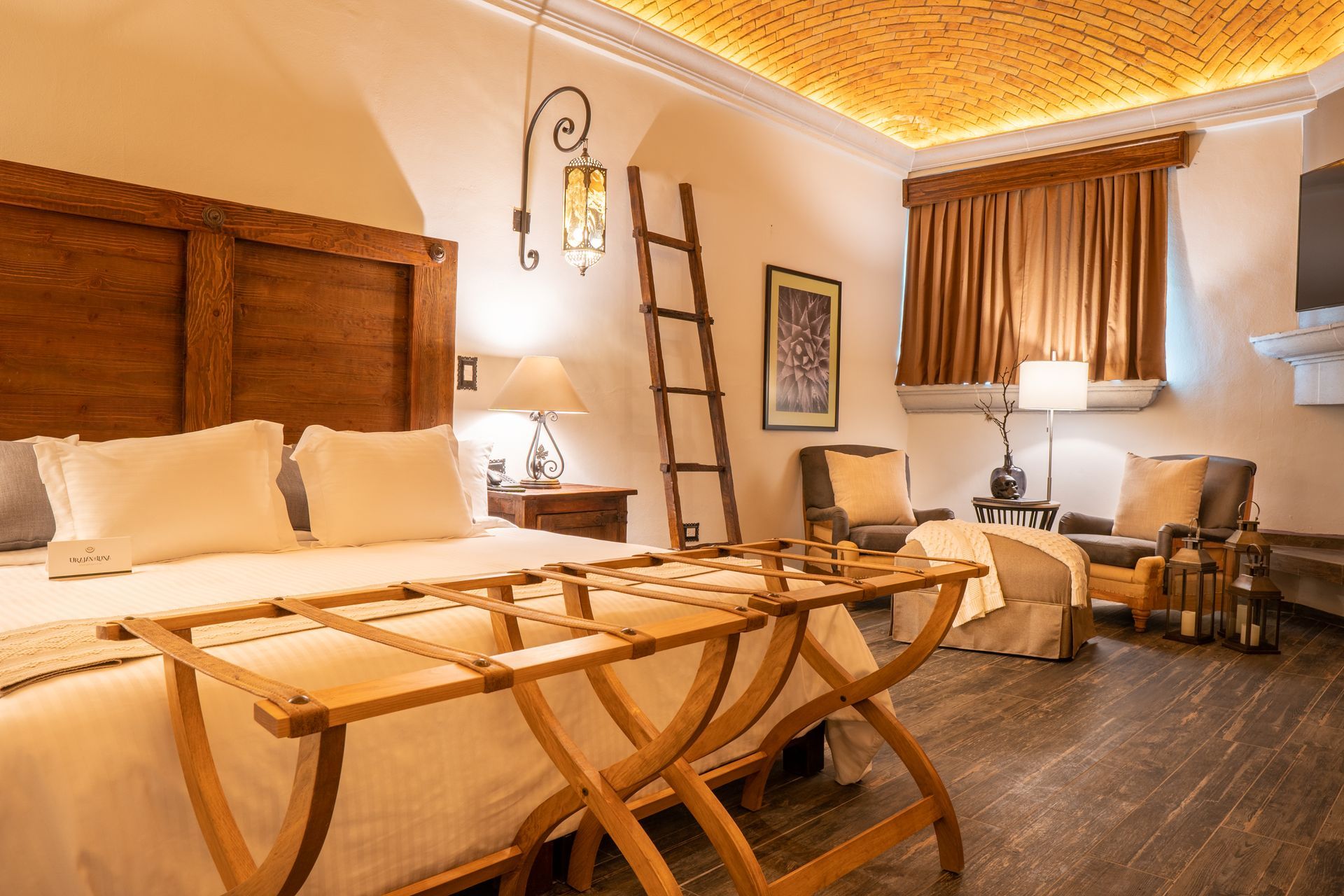 Hotel room with a large bed, seating area, ladder, and wooden luggage rack. Warm lighting, ornate ceiling.