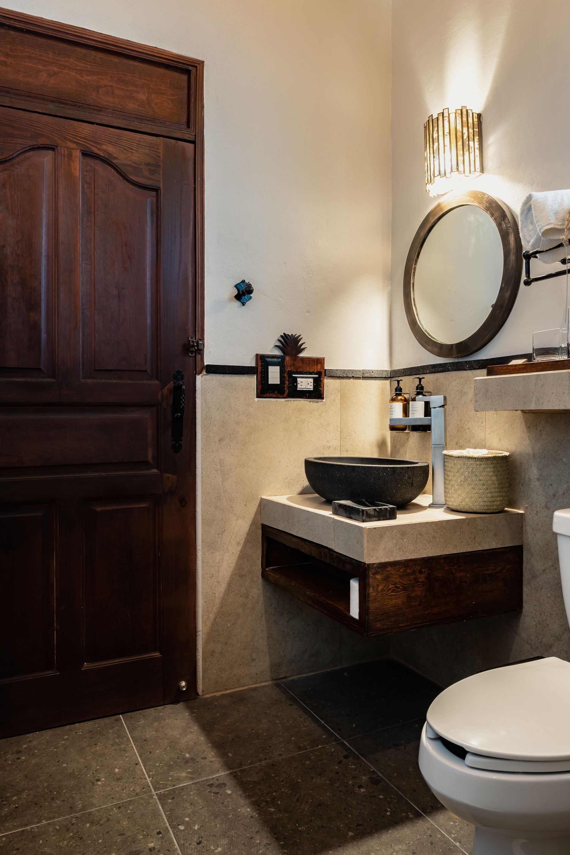 Bathroom with dark wood door, floating vanity with black sink, round mirror, and toilet.