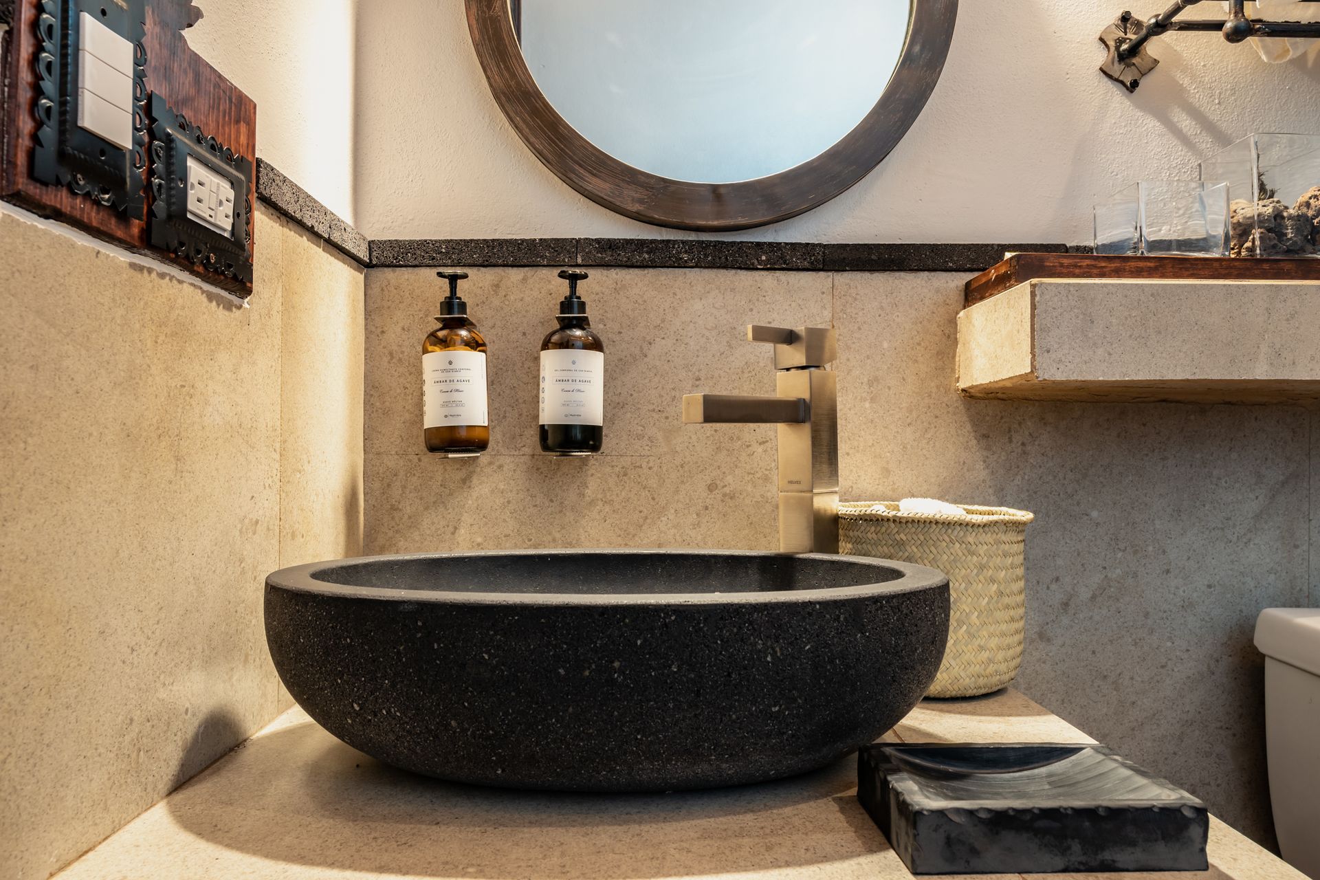 Bathroom sink with a black, textured bowl, a round mirror, and soap dispensers.