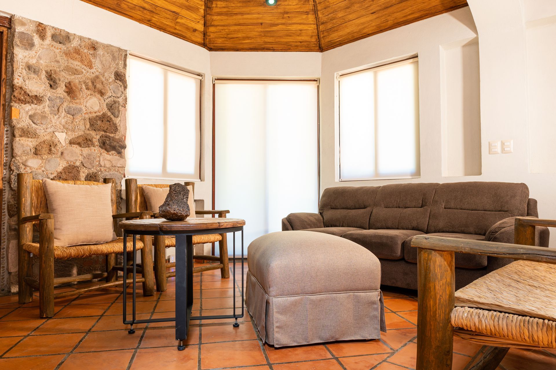Living room with stone wall, wooden furniture, sofa, windows, and a woven straw roof.