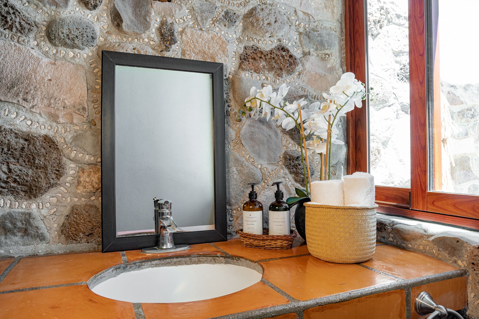 Bathroom sink with mirror, stone wall, window, and toiletries.