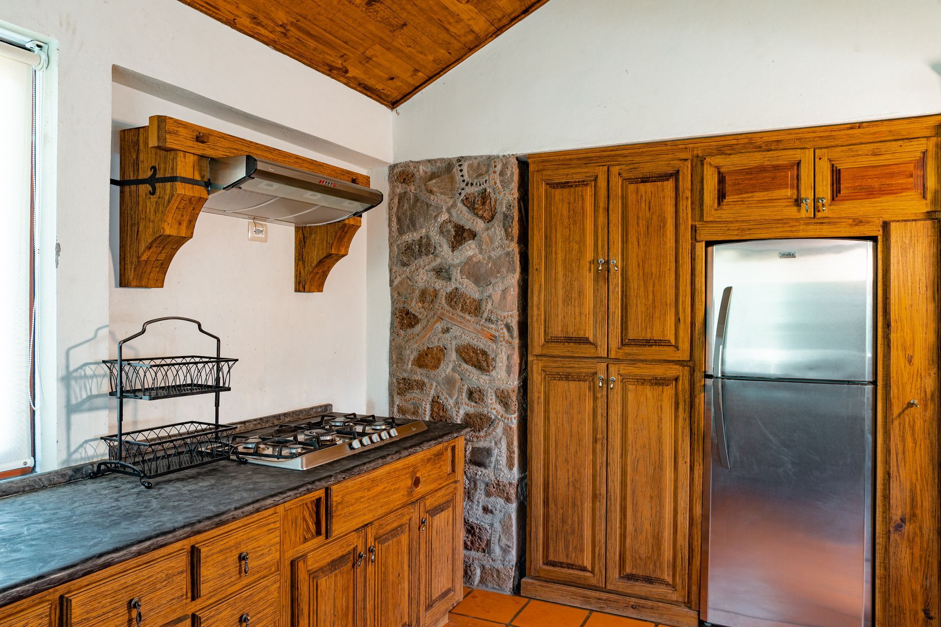 Kitchen with wooden cabinets, stone wall, stainless steel refrigerator, and cooktop.