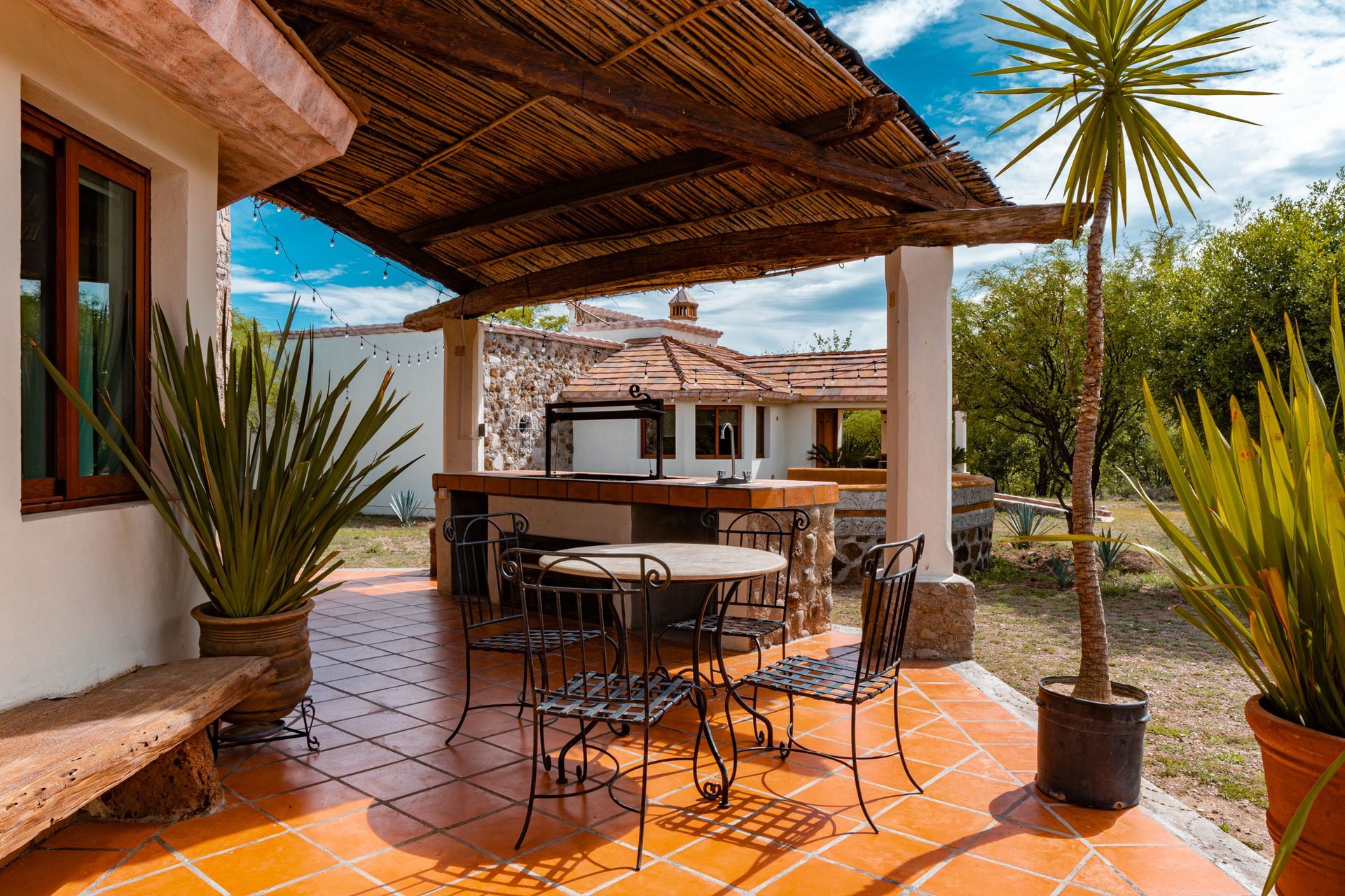 Patio with table and chairs under a thatched roof, outdoor grill in the background, terracotta tile flooring.