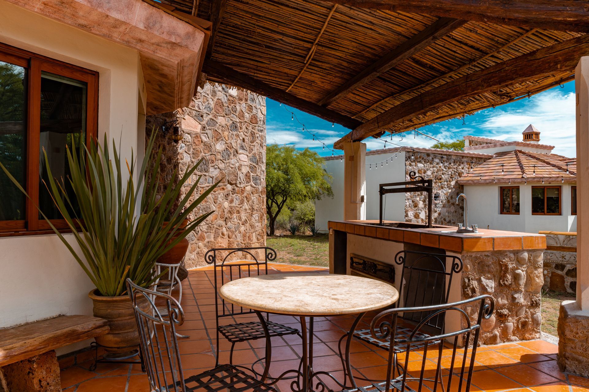 Patio with stone wall, table and chairs, built-in grill, and partial view of a building with a red-tiled roof.