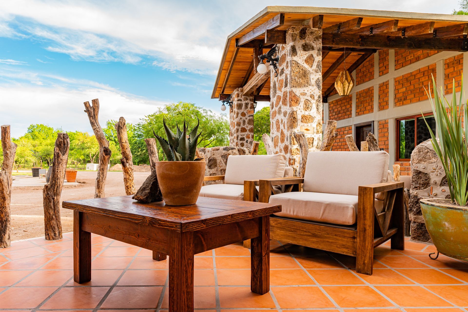 Outdoor patio with wooden furniture and stone columns, tile flooring, plants, and a blue sky.