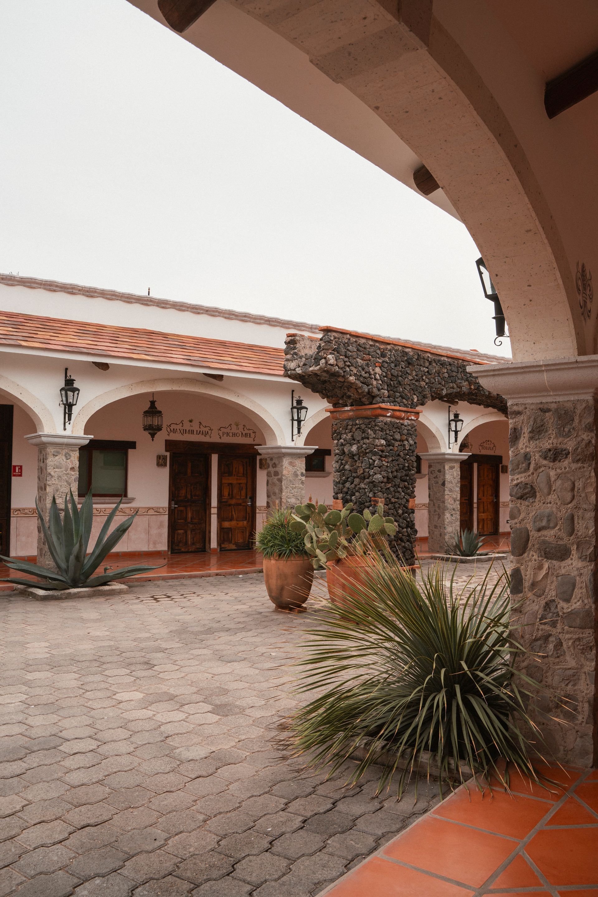 Patio con columnas de piedra, tejado de tejas rojas y plantas. Entrada arqueada.