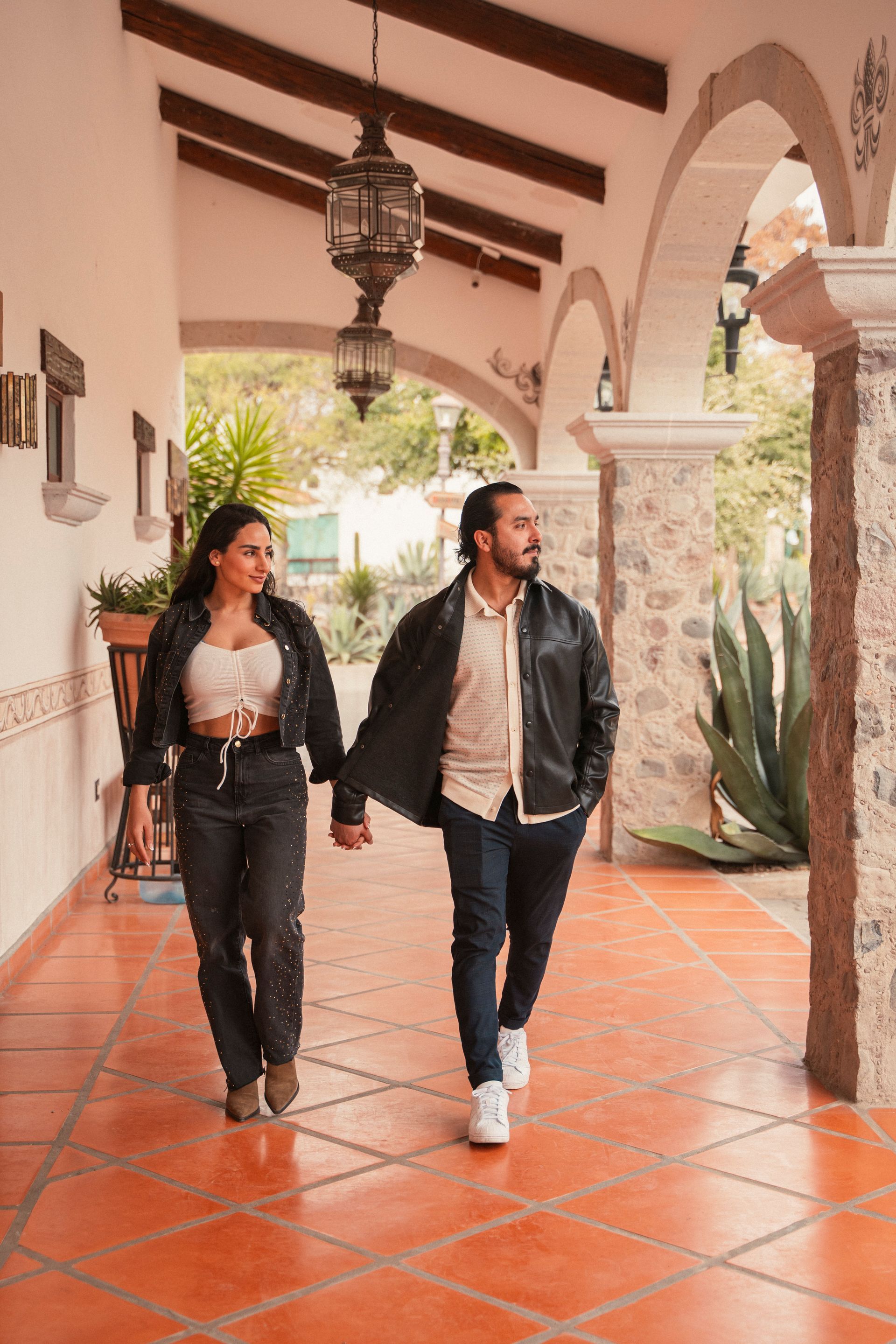 Pareja caminando de la mano bajo un arco de piedra. Suelo de baldosas rojas.