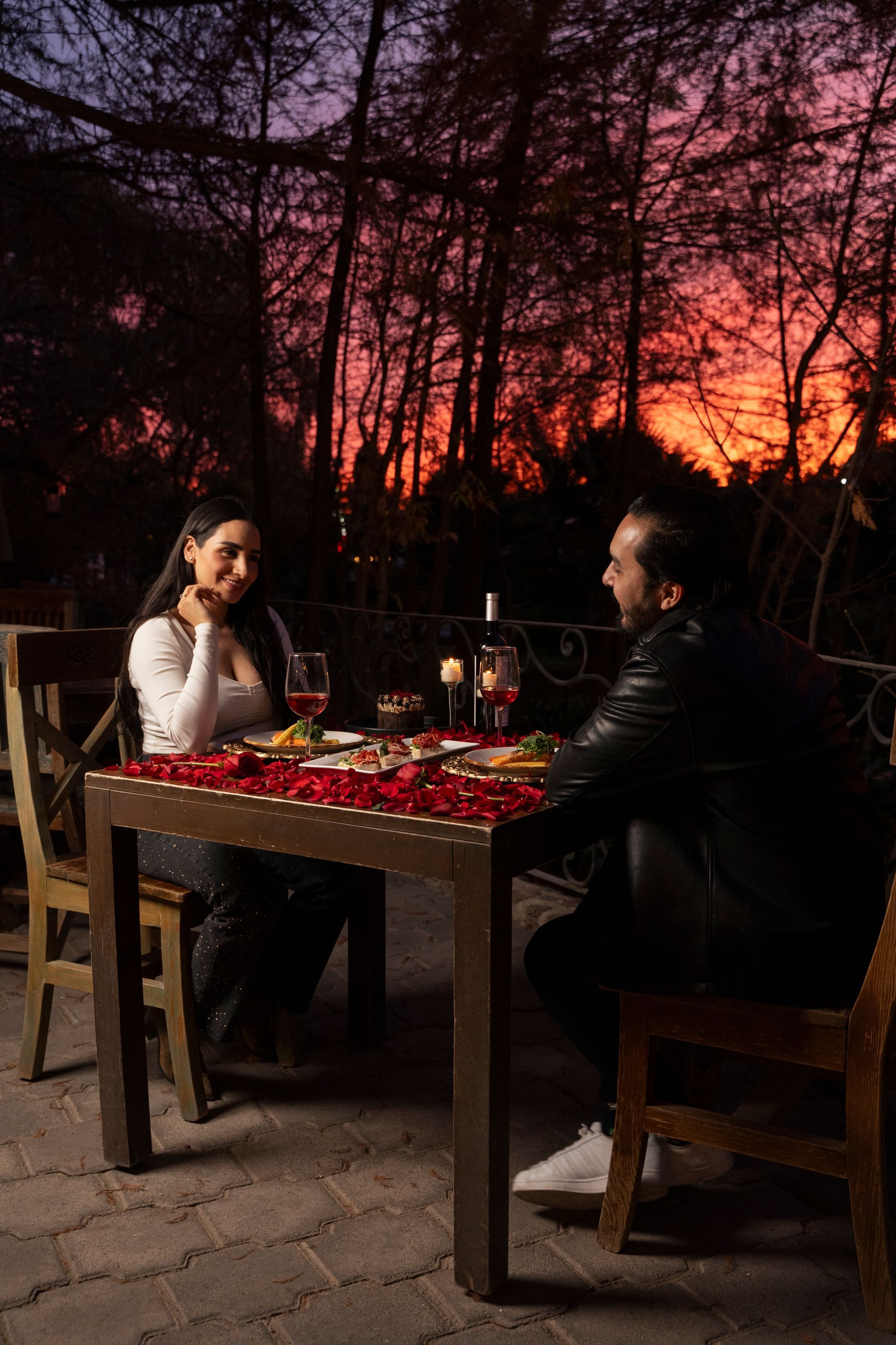Pareja cenando a la luz de las velas, pétalos de rosa sobre la mesa, puesta de sol de fondo.