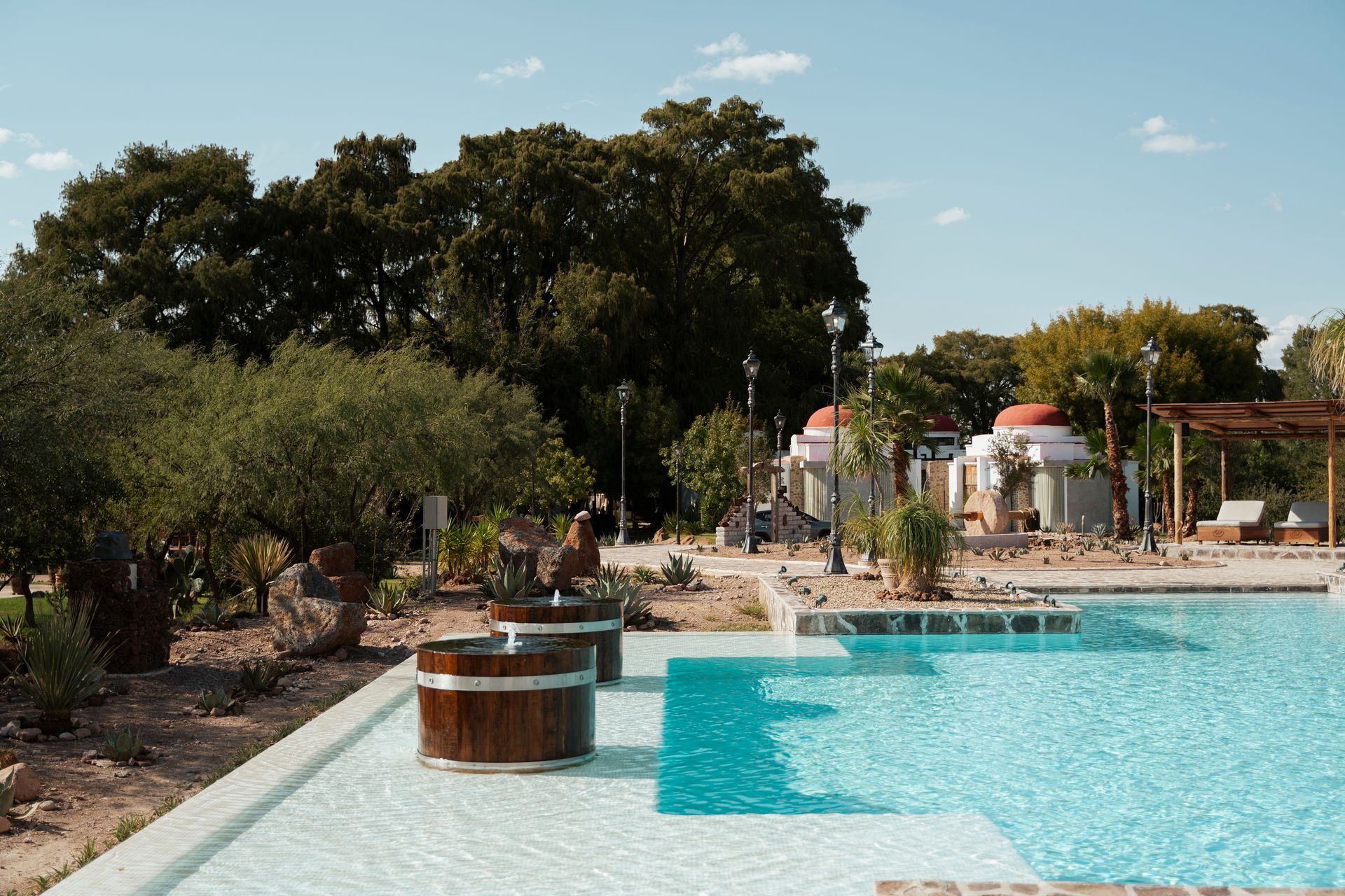 Piscina con bañeras de madera, edificios de spa y exuberante vegetación bajo un cielo brillante.