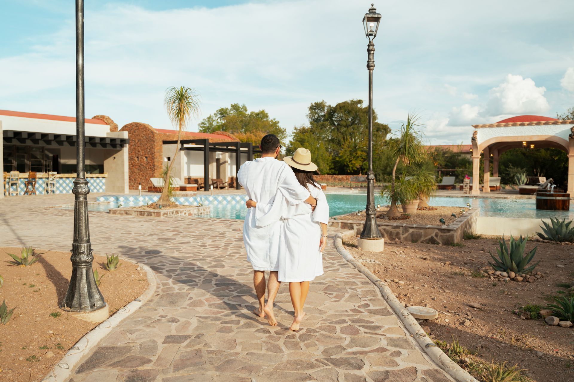 Una pareja con batas blancas se aleja por un sendero de piedra, en dirección a un spa con piscinas y jardines.