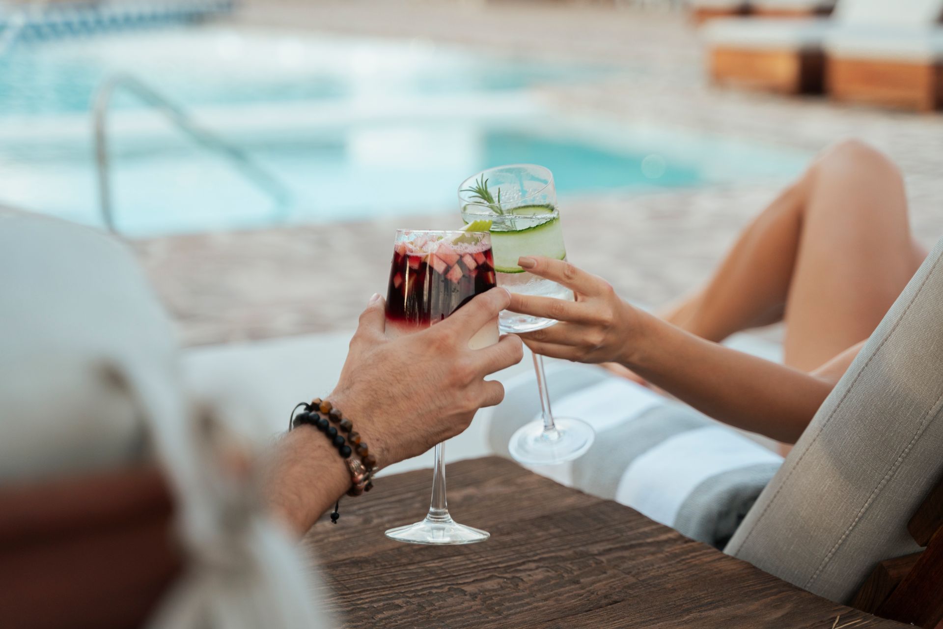 Dos personas brindan con copas de cóctel junto a una piscina. Una sostiene una bebida roja, la otra una verde con pepino.