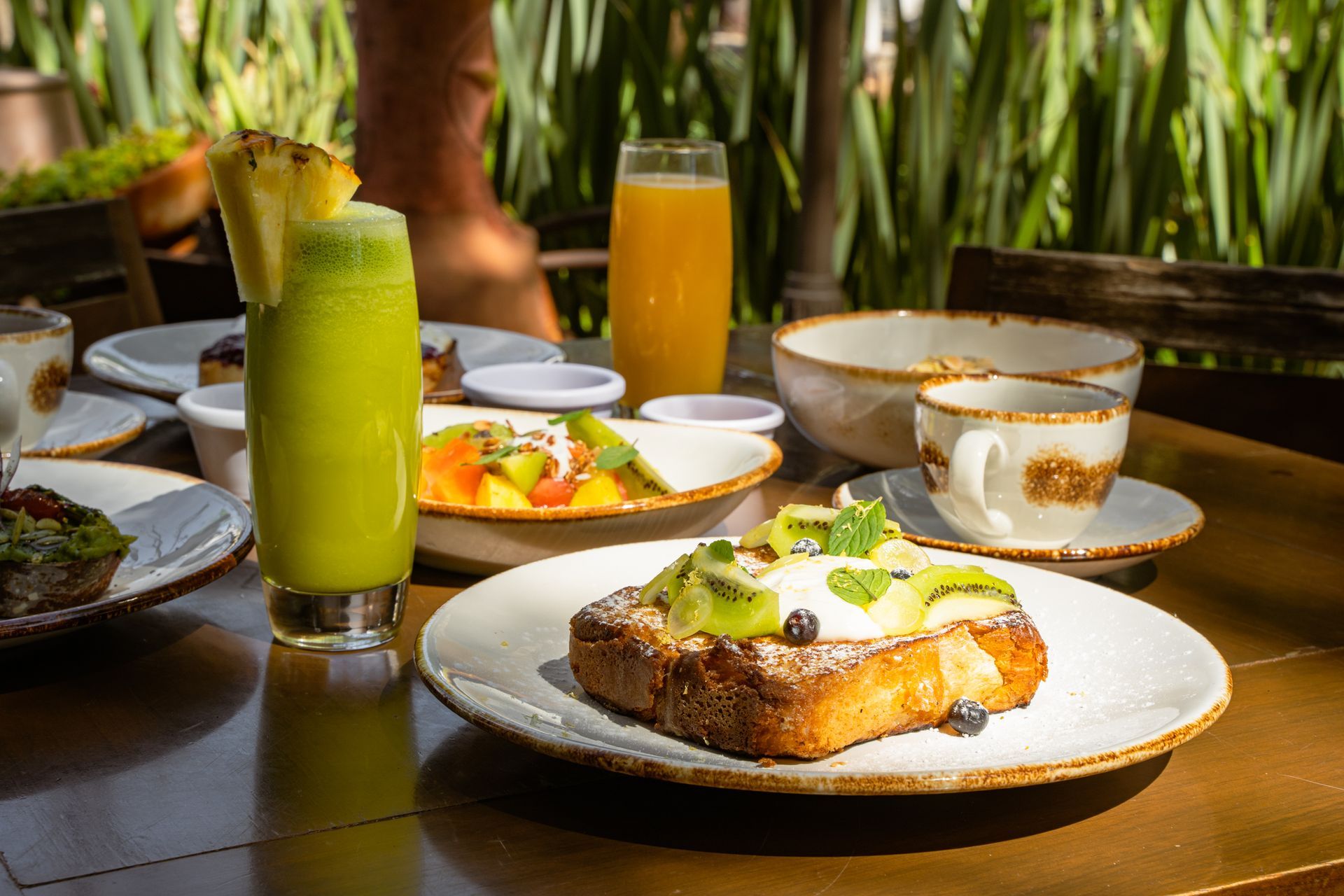 Desayuno completo con tostadas, fruta, zumo y café sobre una mesa de madera al aire libre.