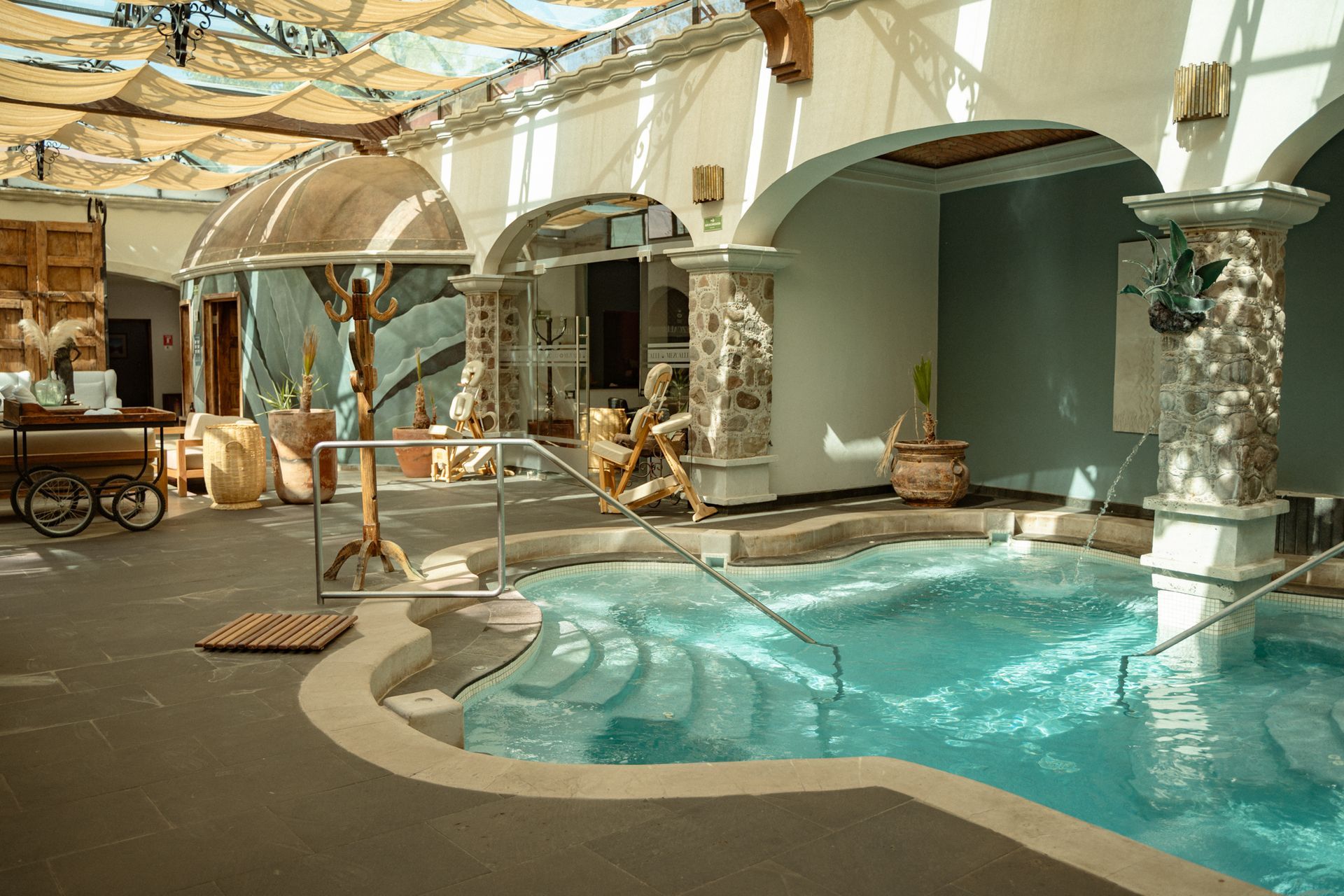 Indoor spa with blue pool, stone columns, arched doorways, and a light canopy roof.