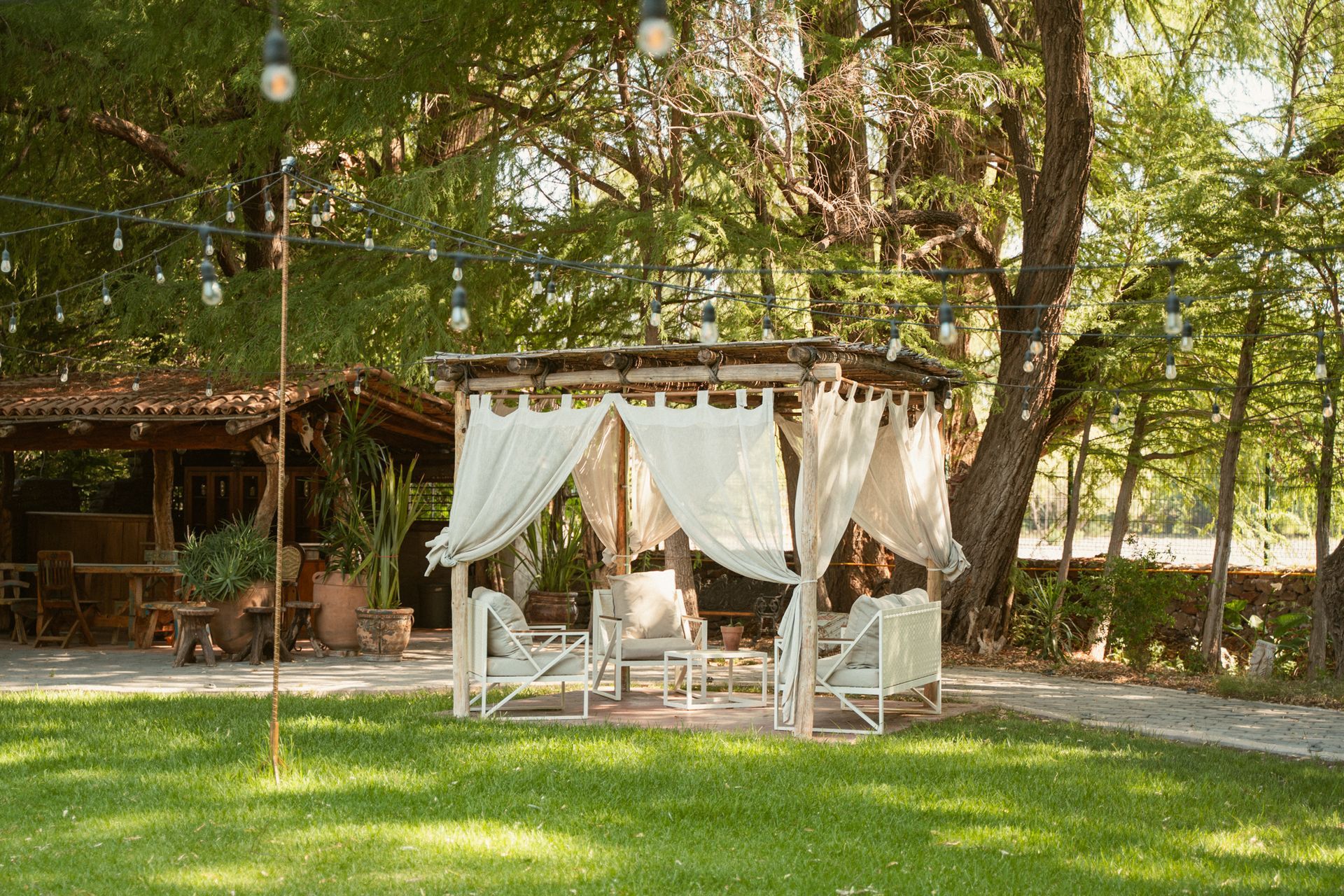 Outdoor seating area with white curtains, chairs, and a table under a wooden pergola in a grassy yard.