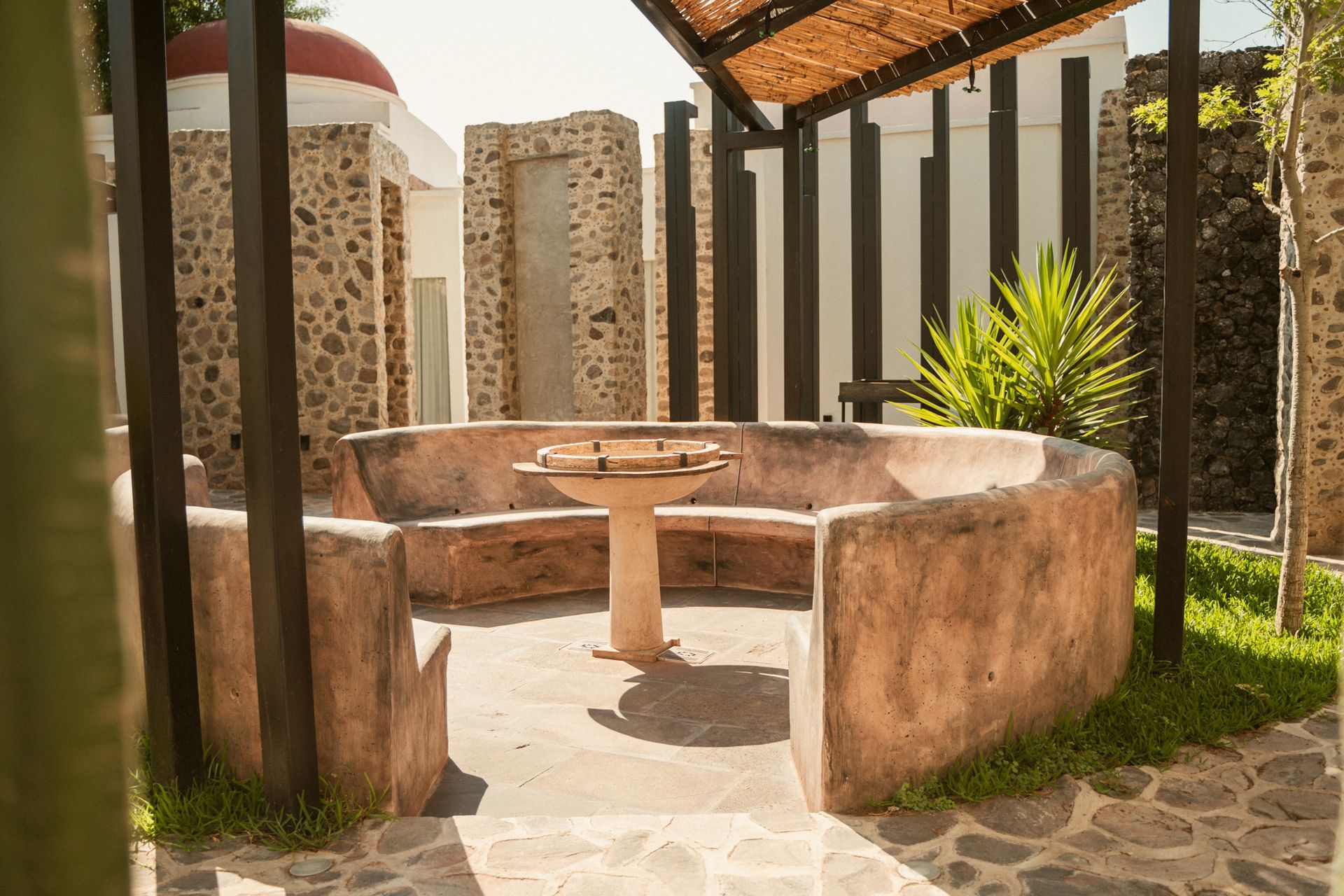Zona de estar al aire libre con un banco circular de hormigón y una parrilla central sobre pedestal.
