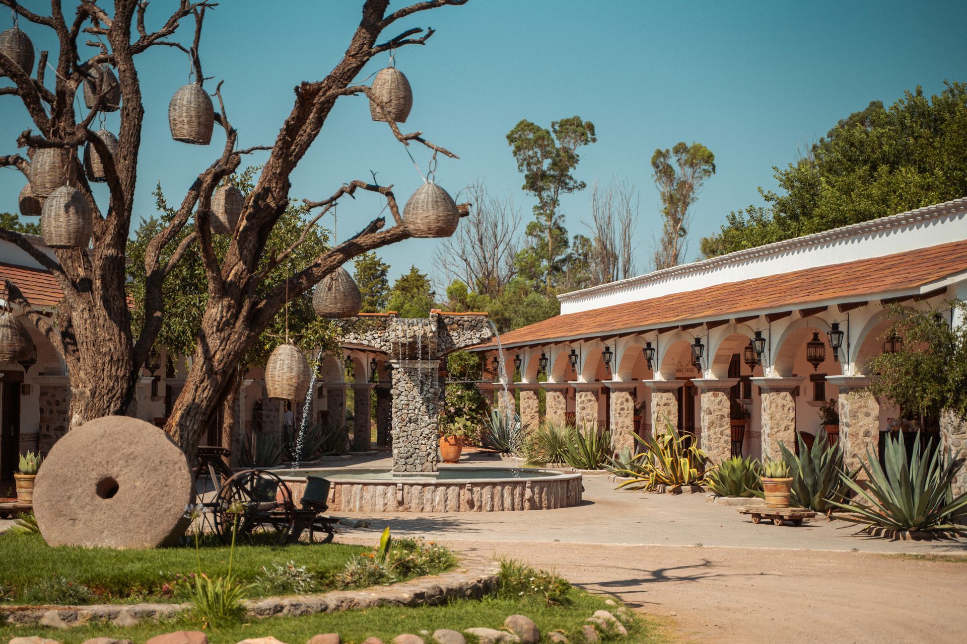 Courtyard with fountain, stone mill, and building with arched columns; decorative tree with hanging woven spheres.