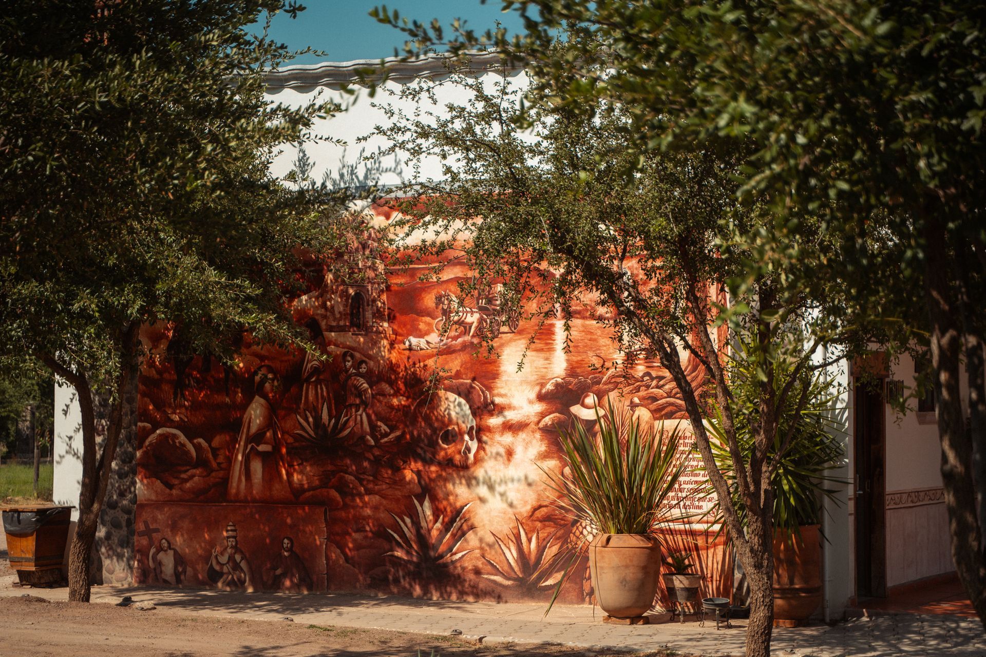 Mural en un edificio blanco, que representa figuras y plantas en tonos naranja y marrón, enmarcado por árboles.